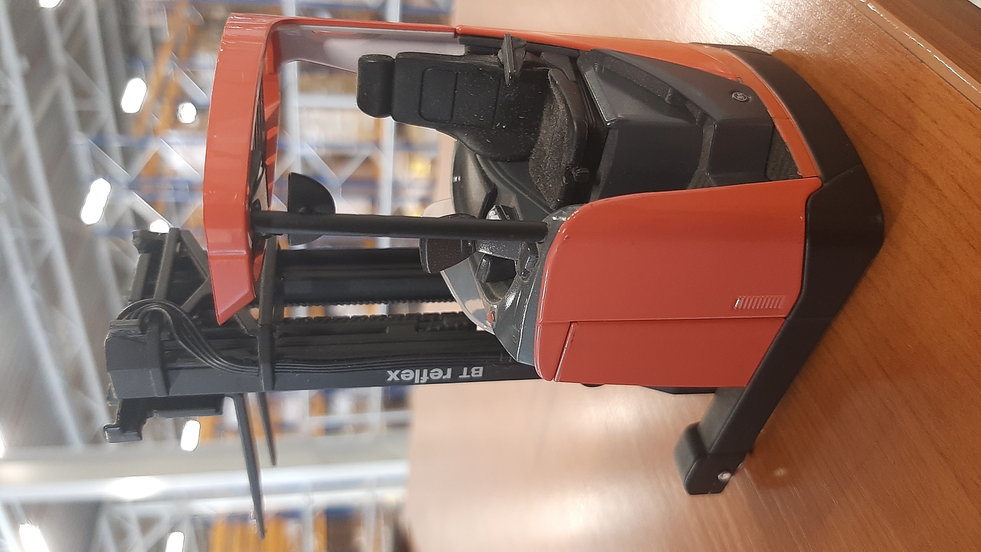 A close-up, eye-level, medium shot captures a red and black model forklift, identified by the "BT reflex" label on its mast, positioned on a wooden table. The forklift, detailed with a driver's seat, control panel, and forks, is the primary subject. The background, slightly out of focus, reveals an indoor environment with high ceilings, industrial shelving, and overhead lighting, suggestive of a warehouse or distribution center. No people are present in the image, and no actions or interactions are depicted. The setting is indoors on a wooden table, with a blurred industrial background. The geographical location of Hellevoetsluis, Netherlands is not discernible from the visual information in the image.
