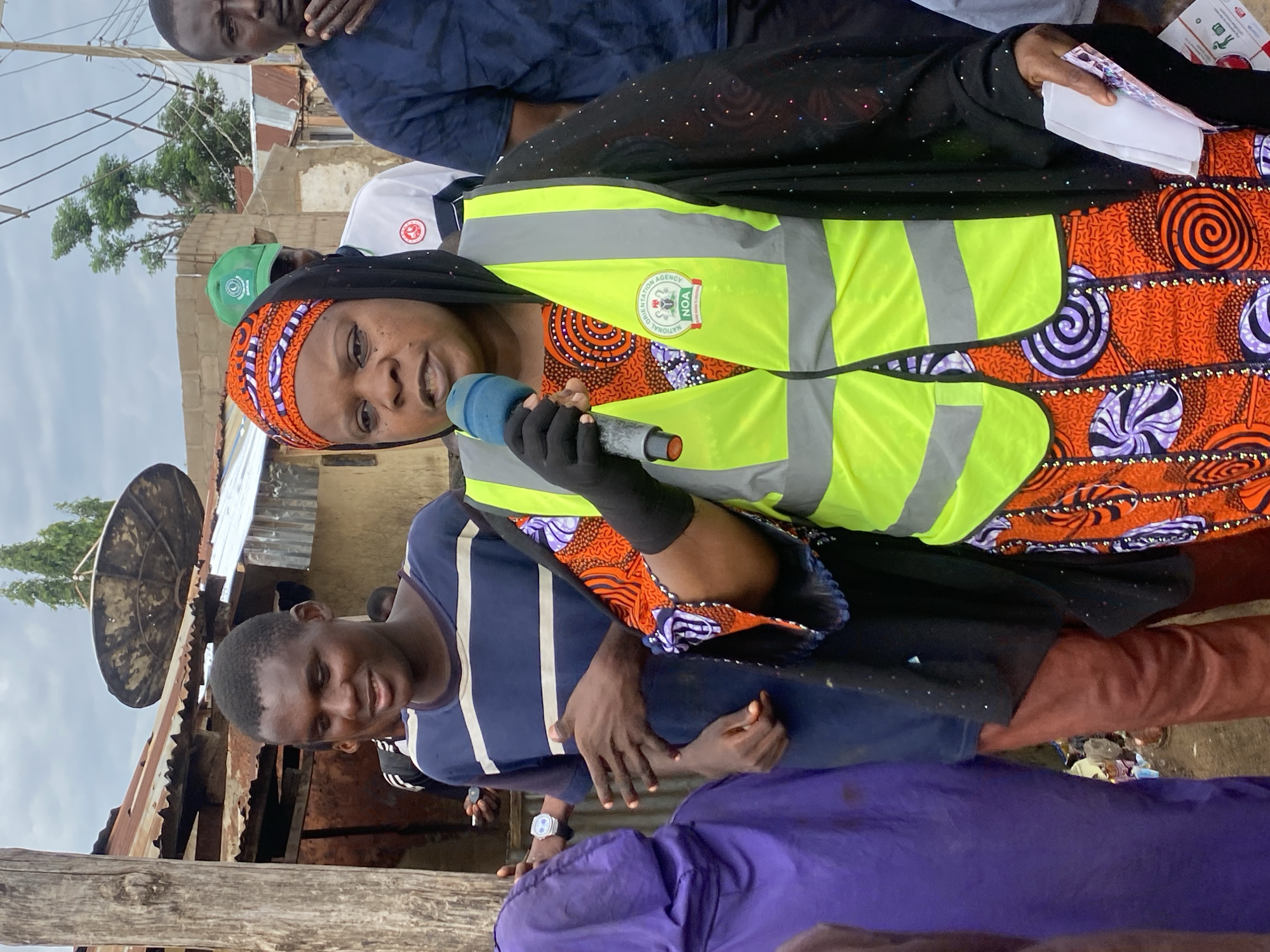 In an outdoor scene in Rigachikun, Nigeria, a woman wearing a black hijab, an orange and purple patterned dress, and a bright yellow safety vest is speaking into a blue microphone. She is holding a stack of white papers in her right hand. The vest has a logo with the words "NATIONAL ORIENTATION AGENCY NOA" printed on it. To her left, a young man in a blue and white striped t-shirt looks towards her, smiling. Behind the woman, another man wearing a green cap and a white shirt is visible. In the background, there are structures with corrugated metal roofs, some appearing worn, and electrical wires running overhead. The ground is uneven and appears to be a dirt path with scattered debris. A purple hooded garment is visible on the far left, suggesting other individuals are present. The overall setting appears to be a modest urban or semi-urban environment.