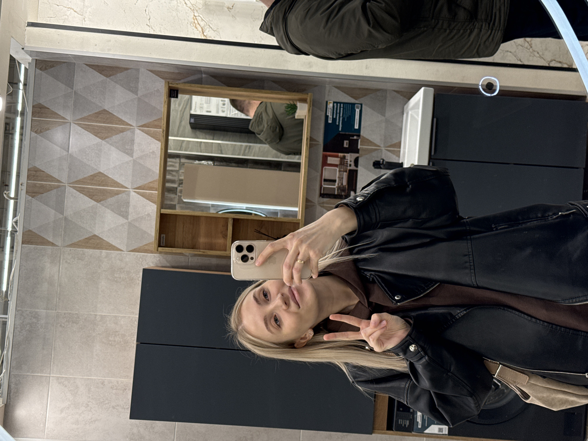 A person takes a selfie in a bathroom with light-colored tiles decorated with geometric patterns and dark grey cabinets, as seen in a reflection from a mirror in the city of Izhevsk, Russia. The person is wearing a black leather jacket over a brown garment and holding a smartphone, making a peace sign. Reflected in the mirror are a man wearing a green jacket and a wooden cabinet. The environment consists of a bathroom with a sink, cabinets, and wall tiles.
