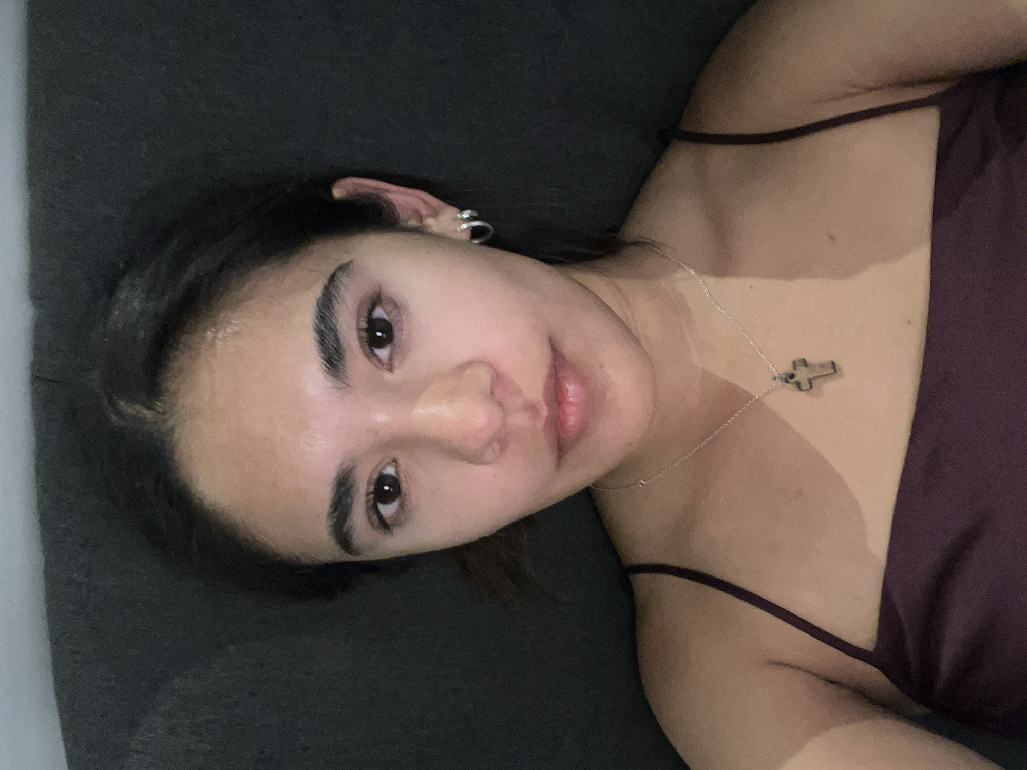 A person with brown hair, wearing a maroon camisole and a silver cross necklace, is lying down on a dark gray surface. They have silver hoops in their left ear. The location is not discernible from the image, and there is no evidence to suggest it is in San Bernardino Tlaxcalancingo, Mexico.