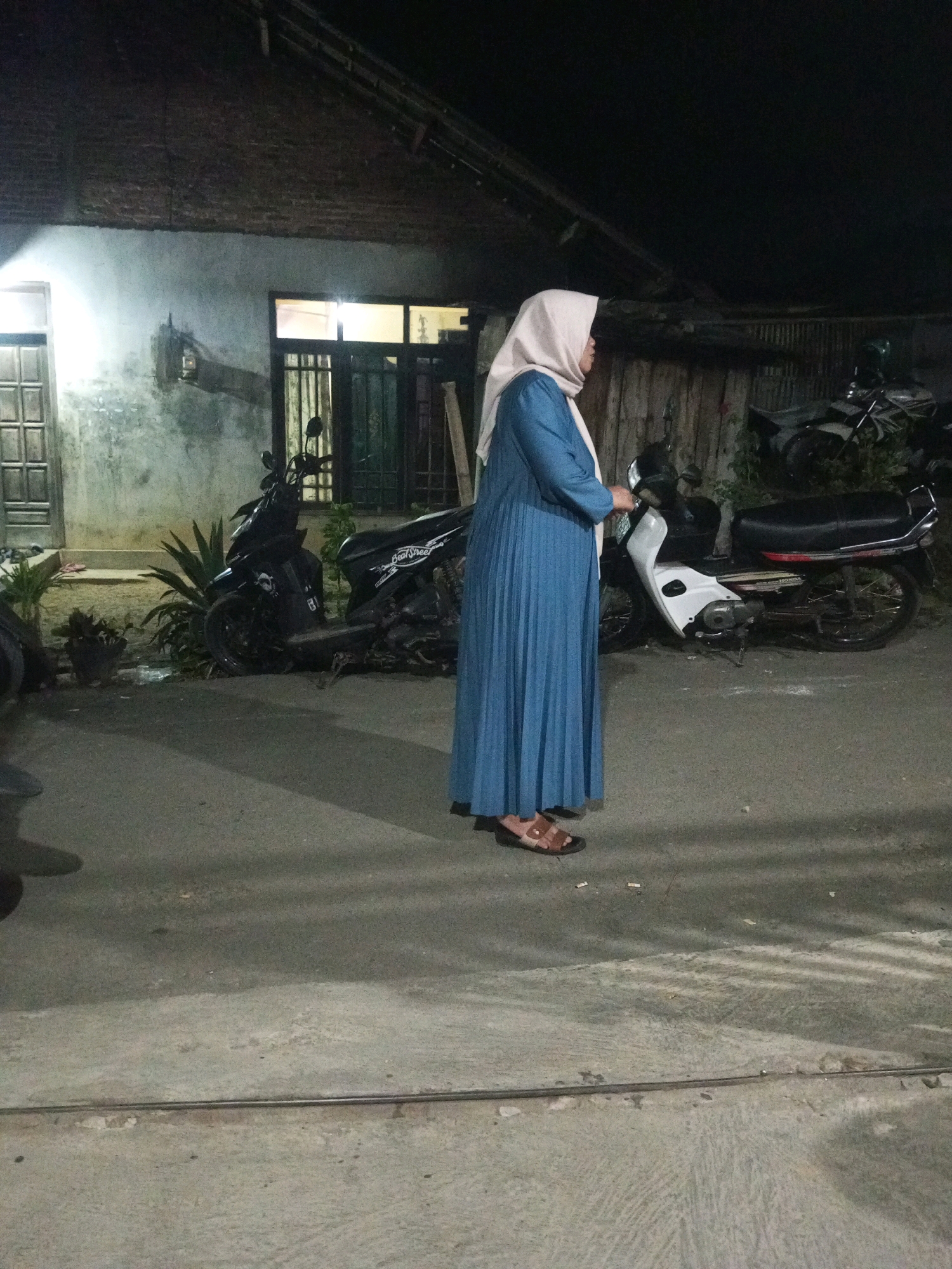 A side profile, medium shot captures a woman wearing a light-colored hijab and a long, blue, pleated dress, standing on a paved surface. She is facing left and her left hand is slightly raised, holding what appears to be a mobile phone. She wears brown sandals. Behind her, parked on the paved surface, are two motorcycles. The motorcycle on the right is white and red, with the brand name "Honda" visible. The motorcycle on the left is dark-colored. In the background, a building with a dark, textured roof and light-colored, weathered walls is visible. The building has windows with dark frames and some with bars. There are plants, including a spiky plant in a pot and some leafy green plants, situated between the building and the motorcycles. The scene appears to be outdoors, possibly in a residential area, and it is nighttime, indicated by the dark sky and artificial lighting. The location is identified as Wonokromo, Indonesia.