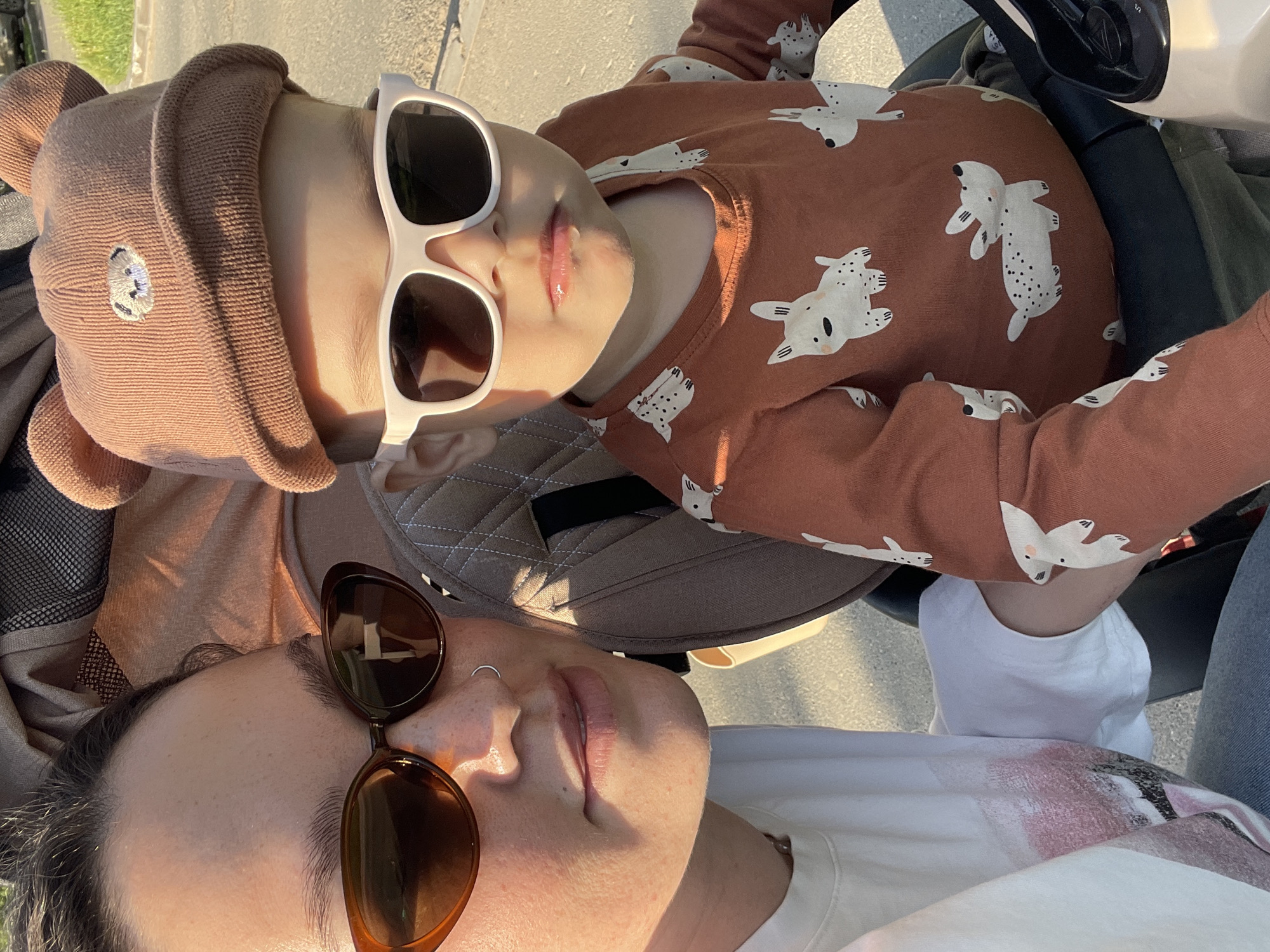 A close-up, angled shot shows an adult and a toddler outdoors. The adult is wearing brown sunglasses and a white t-shirt and has a nose ring. The toddler wears white sunglasses and a brown bear hat. The toddler also wears a brown shirt with dog figures. They are both seated, possibly in a stroller. The location is presumably Izhevsk, Russia, but this is not verifiable from the image itself.