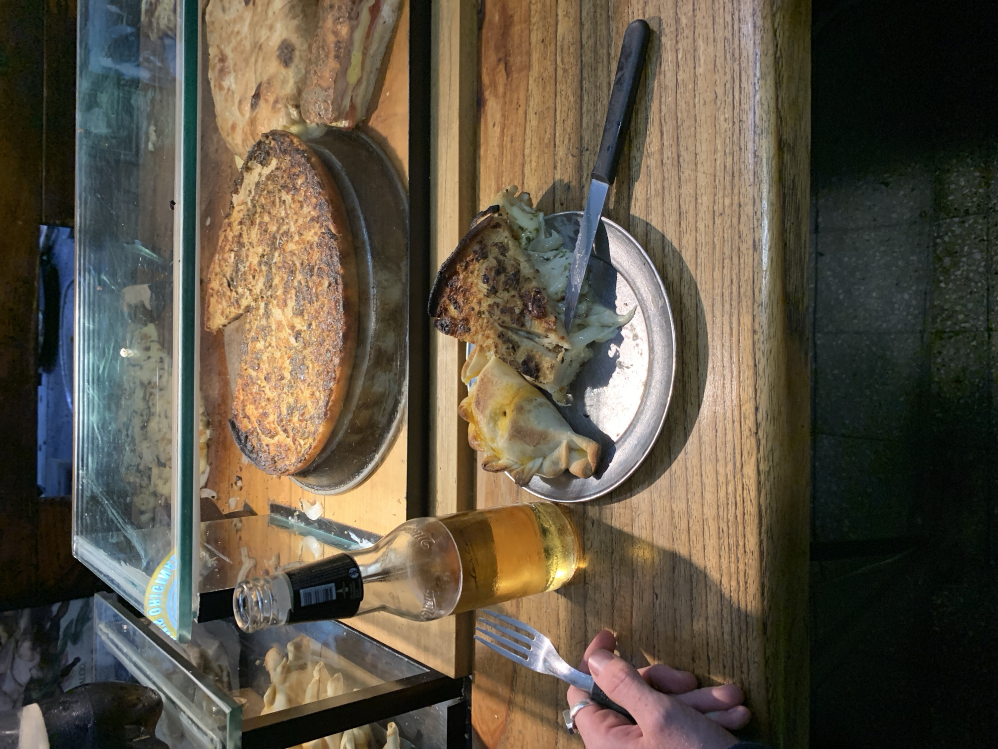The image, taken from a slightly elevated, vertical perspective, captures a casual dining scene on a wooden counter, likely in Buenos Aires, Argentina. The main subjects are various food items, a bottled drink, and a hand holding a fork.
On the left, a large, round, baked dish with a rich, browned, and somewhat charred crust, possibly a savory tart or quiche like a *torta pascualina*, sits in a metal tin. Next to it, partially visible, is another flatbread-like item. These are displayed within or near a glass display case, suggesting a bakery or eatery counter. Another glass display case is visible in the lower left, containing what appear to be fried items or small pastries.
To the right, on a small metal plate, rests a slice of a similar baked dish, showing a pale, possibly onion or potato-based filling, along with an *empanada*. A knife with a black handle is stuck into the slice on the plate. Below the plate, a clear glass bottle containing a golden liquid, likely beer, lies horizontally on the wooden counter. The label on the bottle clearly shows the word "ORIGINAL" in blue lettering, along with a barcode. A person's hand, wearing a ring, holds a silver fork, positioned as if about to eat or having just paused.
The activity depicted is a meal in progress, emphasizing a rustic and unpretentious setting. The lighting suggests indoor artificial light or late afternoon sun, casting distinct shadows from the bottle and plate onto the wooden surface. The wooden counter itself has a warm, textured grain. The floor or a dark background is visible on the far right, composed of small, speckled tiles or concrete. The food items like empanadas and savory tarts strongly suggest the Argentine context provided.