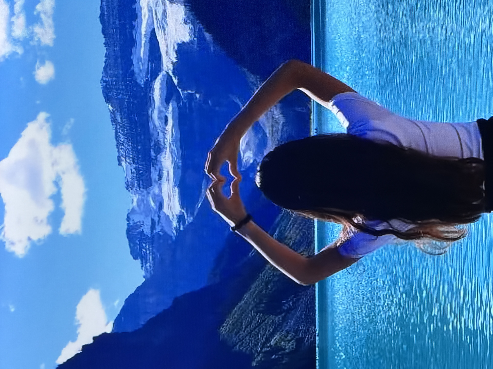 A person stands with their back to the camera, holding their hands up to form a heart shape against a backdrop of a bright blue sky, rugged mountains capped with snow, and a turquoise lake. The person has long, dark hair and is wearing a white t-shirt with thin horizontal stripes and a black bracelet on their left wrist. The lake's surface is rippled, reflecting the light. The mountains are steep and imposing, with visible geological textures and snow-covered peaks. The sky is clear except for a few fluffy white clouds. The overall scene conveys a sense of natural beauty and appreciation.