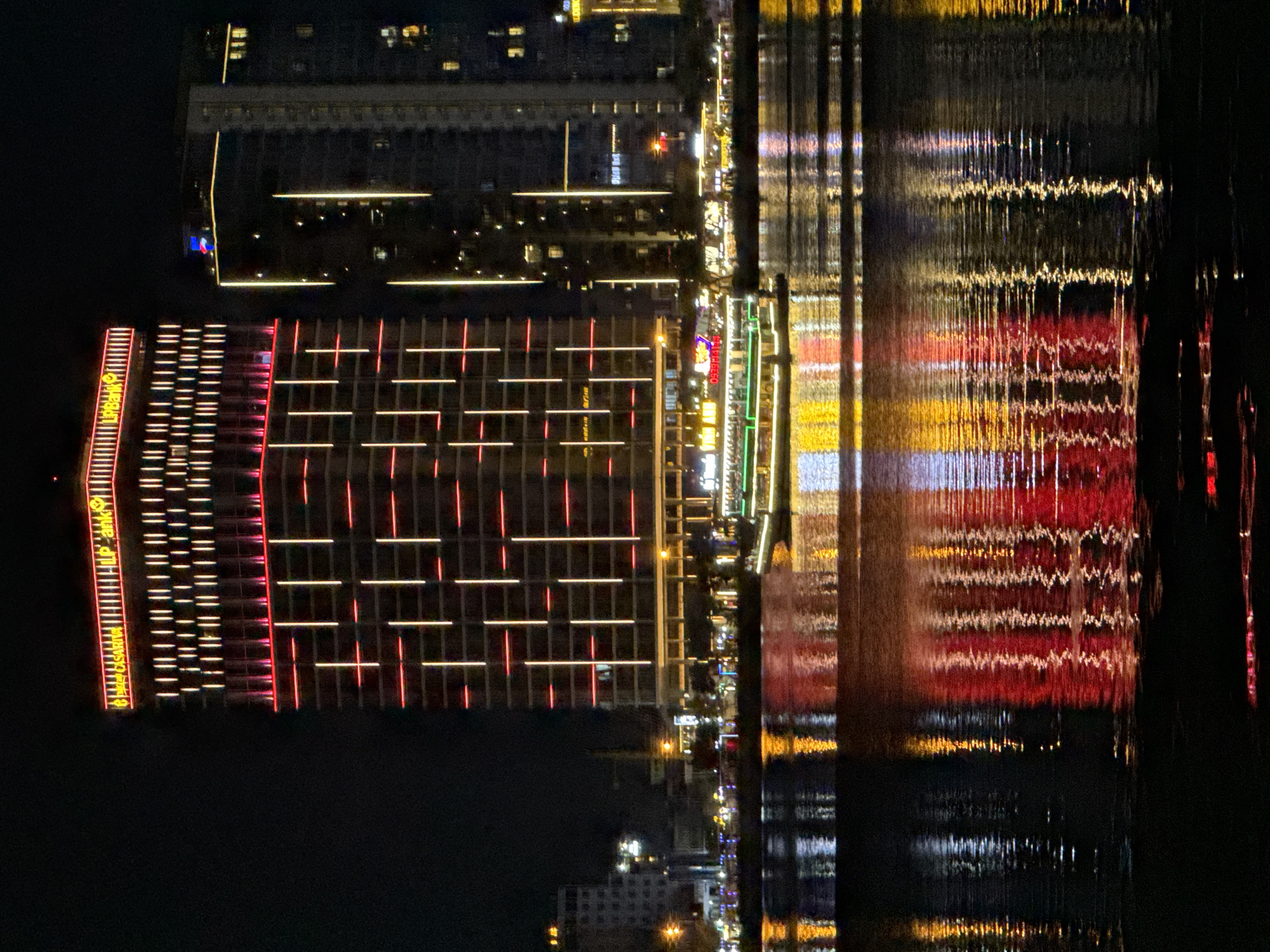 This nighttime scene depicts the illuminated skyline of Đà Nẵng, Vietnam, reflected in the calm waters of a river or bay. The distinct building on the left features a grid-like pattern of horizontal white and vertical red lights, and signage for "HLP Bank" vertically stacked in yellow. Adjacent to this building, another structure is partially visible with linear yellow lights. A street or promenade runs along the waterfront, illuminated by streetlights, with a mix of yellow and green signage reflected in the water. The reflections create blurred vertical streaks of color (red, yellow, white) in the water. The sky is dark. Other buildings are visible in the background, lit with various lights. The image appears to be taken at night, from a slightly elevated position.