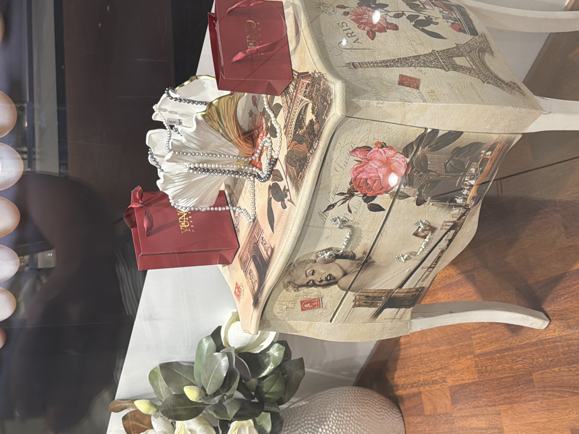 The image shows a decorated side table with drawers, on which are placed two red gift bags, a decorative white shell-like object with pearl necklaces draped over it, and a vase with artificial flowers. The side table itself is decorated with images of Marilyn Monroe, roses, the Eiffel Tower, and other Parisian-themed graphics, including stamps and cursive writing. The table has ornate drawer pulls. The floor beneath is polished wood. The scene appears to be a display in a shop or store. The exact location in the city of Sofia, Bulgaria cannot be determined from the image alone.