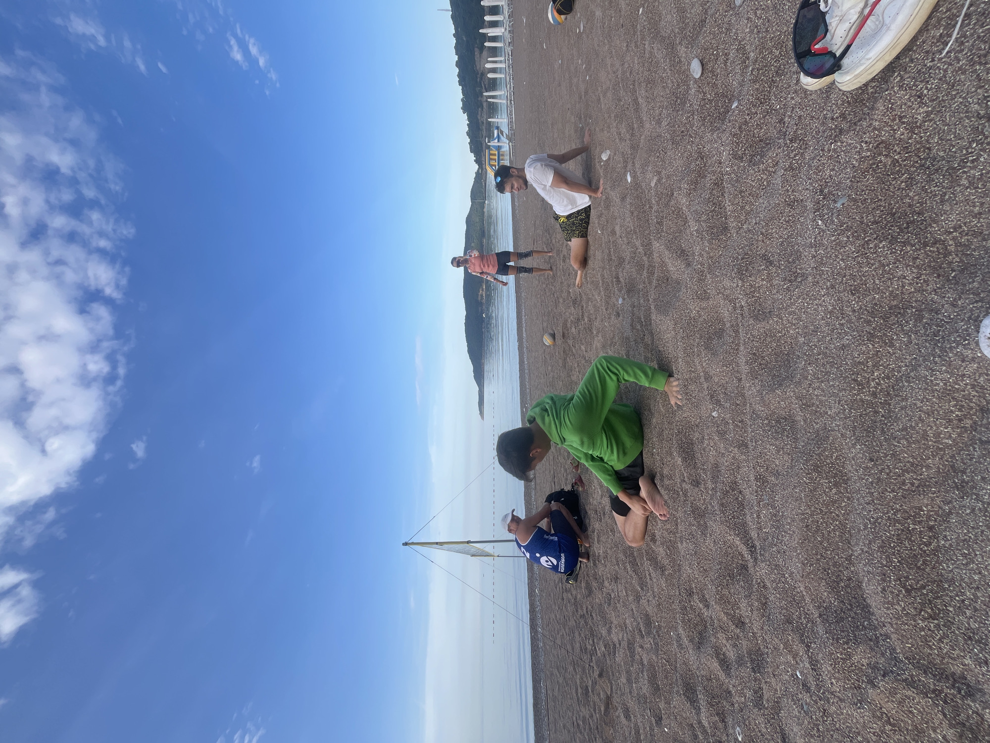 On a sunny beach near Budva, Montenegro, several people are present. Two children are near a small, improvised sailboat, one wearing a blue and white sports shirt and the other in a green sweatshirt. Further along the beach, an adult in a white shirt and patterned shorts is doing push-ups, while another individual in an orange shirt and dark shorts is standing nearby. The beach itself is made of dark sand, and the sea is visible in the background, with a coastal town lining the shore. A pair of white sneakers and sunglasses are also on the sand, away from the people.