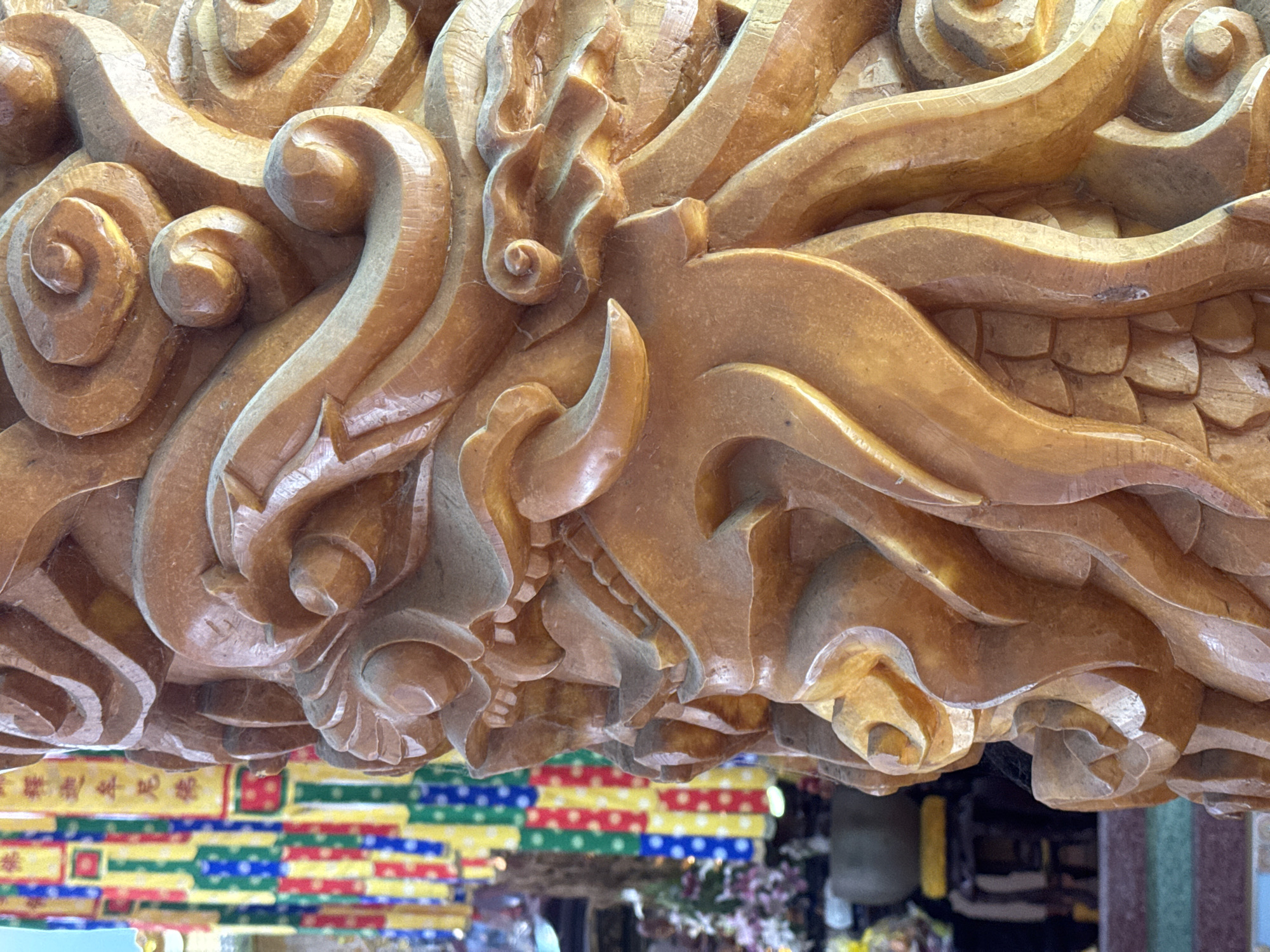 A close-up, low-angle shot features an intricately carved wooden structure. The carving exhibits a complex design with swirling patterns and stylized shapes. The wood has a smooth, golden-brown finish, highlighting the details of the carving. In the background, a colorful display is visible. The display contains rectangles and squares, using yellow, green, blue, and red. The reflection in the surface reveals a glimpse of the interior, with what appears to be floral arrangements and other decorative elements. The scene is in Nha Trang Ward, Vietnam.