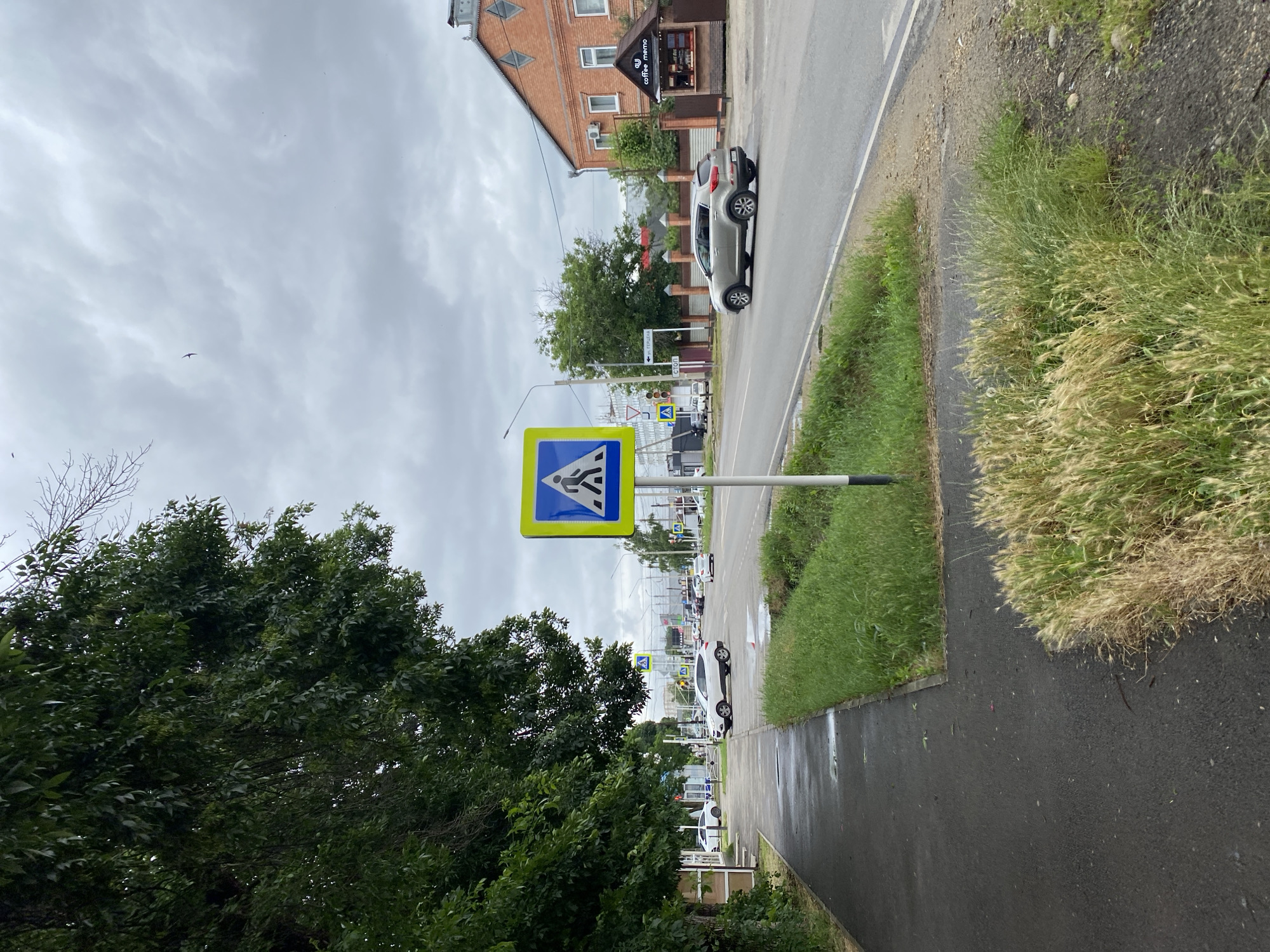 The image shows a street scene in the city of Zapadny, Russia, on a cloudy day. A pedestrian crossing sign is prominently displayed on the right side of the road, featuring a blue square with a yellow border and a black silhouette of a person walking. The sign is mounted on a metal pole. To the left of the sign, a sidewalk is visible, wet from recent rain, with some puddles. Lush green grass lines the edge of the sidewalk and separates it from the road.
The road itself is paved and has at least two lanes of traffic. Several cars are present. A silver SUV is driving on the right side of the road, moving away from the camera. In the background, on the left side of the road, more cars are visible, including a white SUV that appears to be stopped or moving slowly. Further in the distance, traffic lights and other street signs can be seen, along with buildings. One building on the right side of the road is made of red brick and has a small, open-air kiosk with the text "coffee memo" on it. Trees with dark green foliage line the left side of the sidewalk, creating a canopy effect over parts of the scene. The sky is overcast with gray clouds. There are no people clearly visible in the immediate foreground, but there might be pedestrians further down the road near the white SUV.