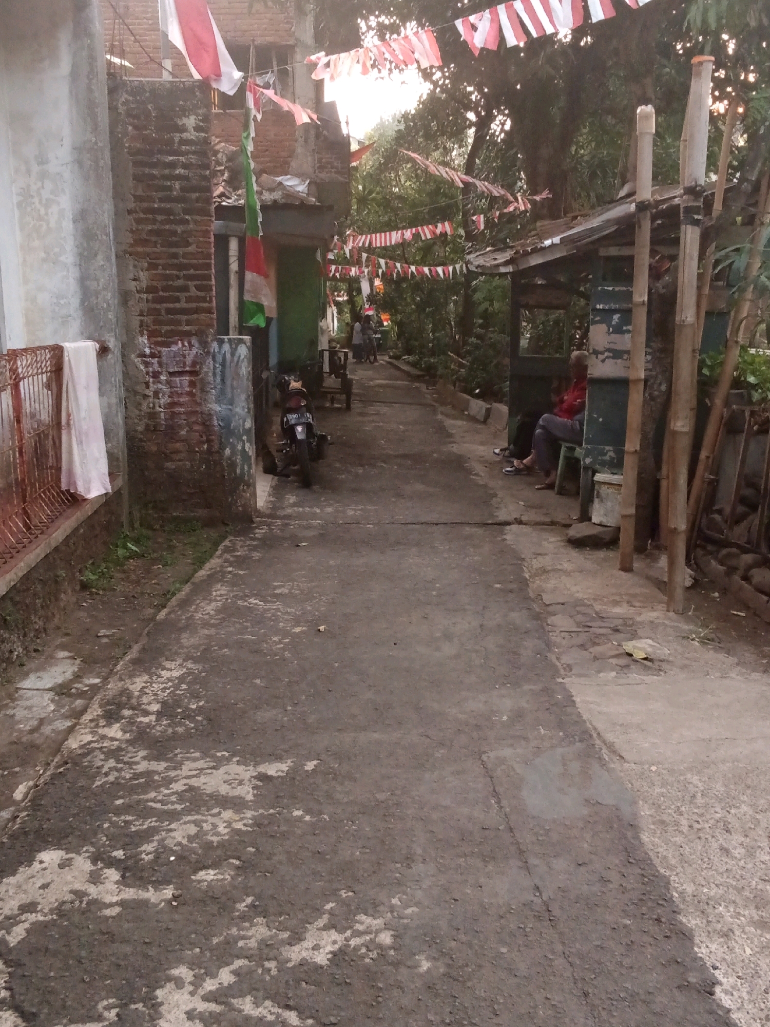 This is a vertical, medium-angle shot of a narrow, paved alleyway in a residential area, likely in Bandung City, Indonesia, given the presence of Indonesian flags. The alley is lined with buildings on both sides, some appearing to be under construction or in disrepair, and dense foliage.
On the left, a weathered, light-colored concrete wall dominates the foreground, with a rusty red metal fence running along its base. A white towel hangs on the fence. Beyond the fence, the ground is covered with dirt and some patchy vegetation.
The alley itself is paved with asphalt, showing signs of wear and tear with lighter patches of concrete exposed. In the middle ground, a dark-colored motorcycle is parked on the left side of the alley.
To the right, a simple, open-sided structure with a corrugated metal roof is supported by bamboo poles. Two people are seated under this structure. One person, wearing a red shirt and blue pants, is facing away from the camera. Another person, wearing dark clothing and sandals, is sitting to their left. A white bucket is placed on the ground near them. A pile of logs is visible behind the structure.
Numerous red and white striped flags, possibly national flags, are strung across the alleyway, suggesting a celebratory or commemorative event. The lighting suggests it is late afternoon or early morning, with soft sunlight filtering through the trees. The overall impression is of a lived-in, somewhat rustic urban environment.