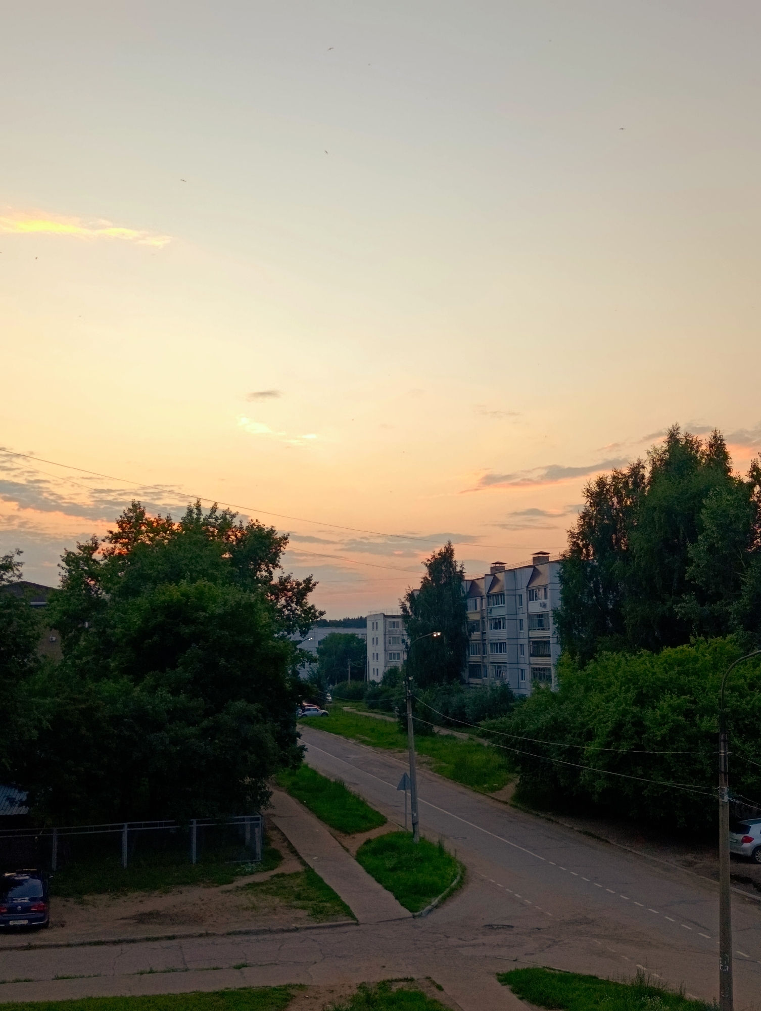 This media file captures an exterior view of a residential area in Chaikovsky, Russia, during what appears to be sunset. The sky is dominated by warm hues of orange and yellow, with streaks of pink and light clouds suggesting a pleasant evening. A few small dark specks, possibly birds, are visible against the vast sky.
The scene depicts a paved road curving through a neighborhood lined with apartment buildings and abundant greenery. Tall, lush trees, some with leaves tinged by the sunset light, are prominent. In the mid-ground, a multi-story apartment building with a light-colored facade and numerous windows stands out. Another apartment building is partially visible on the left.
On the ground level, a sidewalk runs alongside the road, with patches of grass and some dirt areas. A dark blue car is parked near a fence on the left side of the image. Further down the road, another car, a light-colored sedan, is visible on the right. Streetlights and utility poles are also present, with electrical wires crisscrossing the sky.
The overall impression is one of a quiet, residential evening in a seemingly suburban or urban setting. The lighting suggests the transition from day to night, creating a peaceful and serene atmosphere. No text is visible in the image.