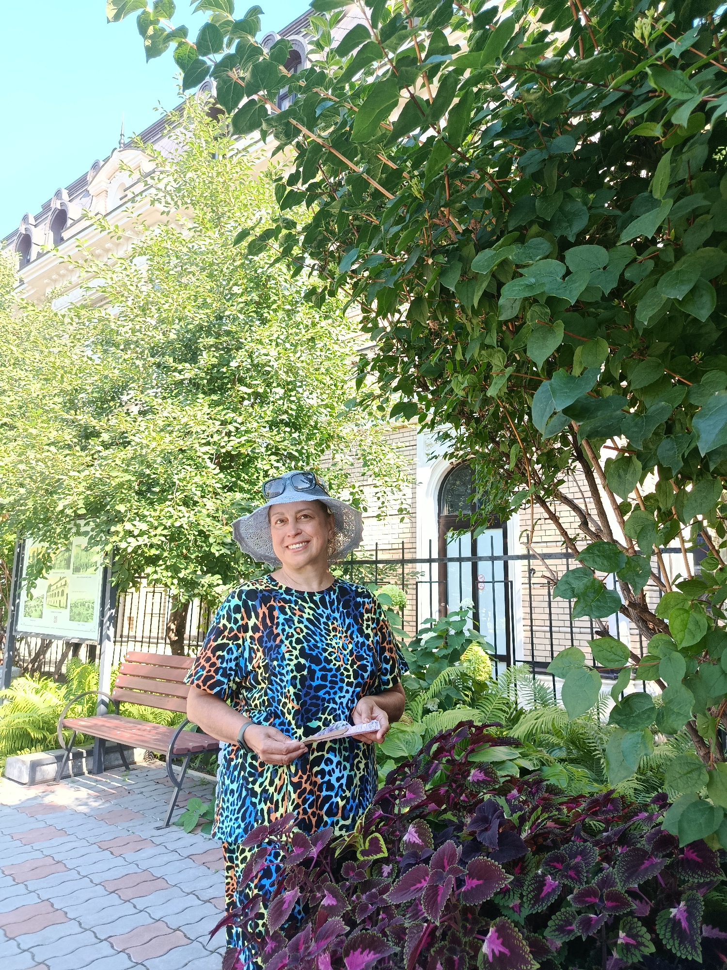 A woman with short, dark hair and sunglasses on her head stands in front of a building in Saratov, Russia, on a sunny day. She is wearing a colorful, leopard-print shirt and a wide-brimmed hat. She is holding a folded piece of paper. To her left is a park bench, a sign with text and images, and lush green foliage. The building behind her appears to be made of brick and has arched windows. The ground is paved with interlocking stones.
