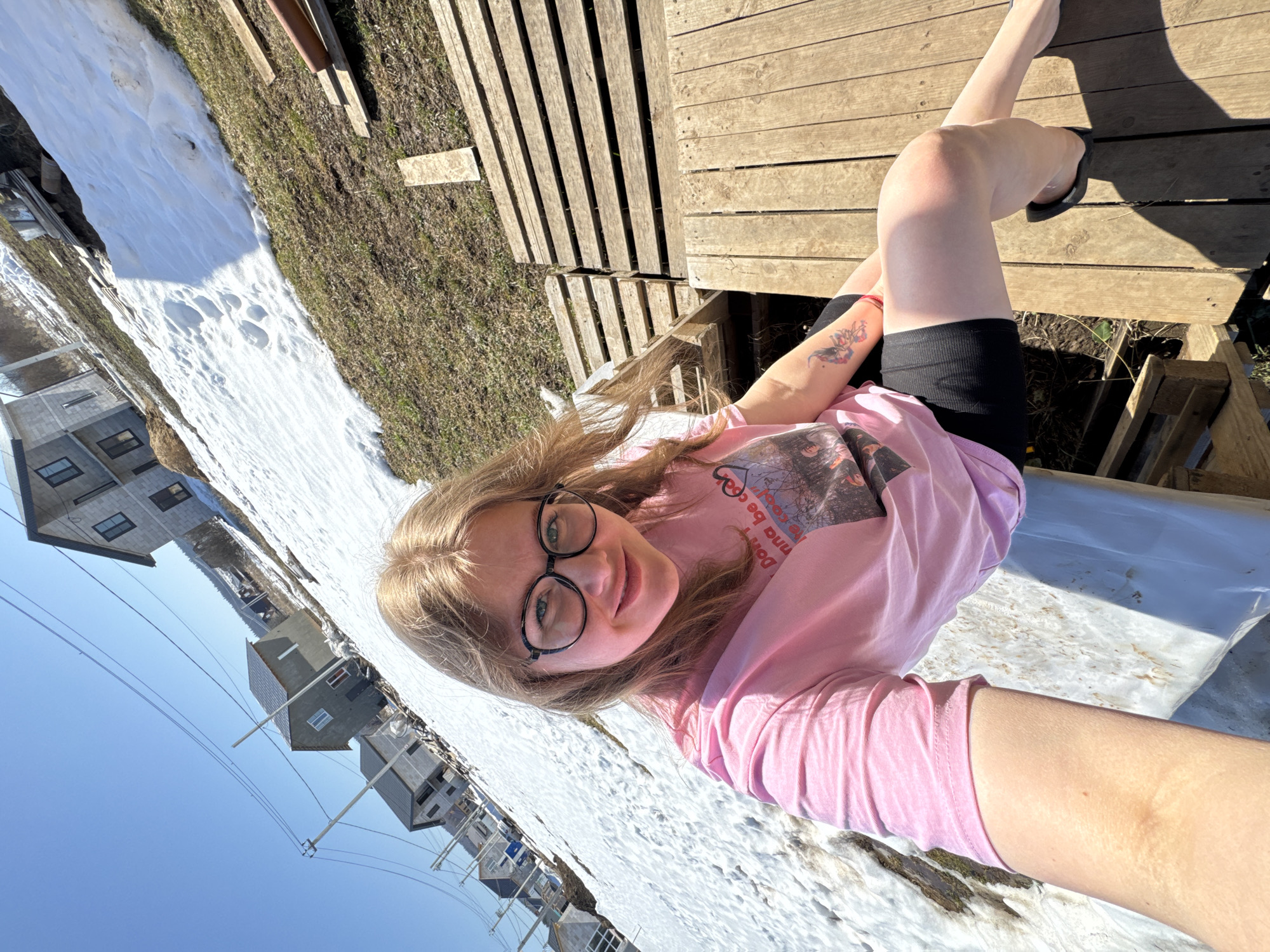 A fair-skinned woman with long blonde hair and eyeglasses sits outside on a wooden deck. She wears a pink t-shirt, black shorts, and black flip-flops, and has a tattoo on her left arm. Snow covers the ground near residential buildings under a clear blue sky. The location is Gornyi, Dvurechenskoe S/P, Russia.