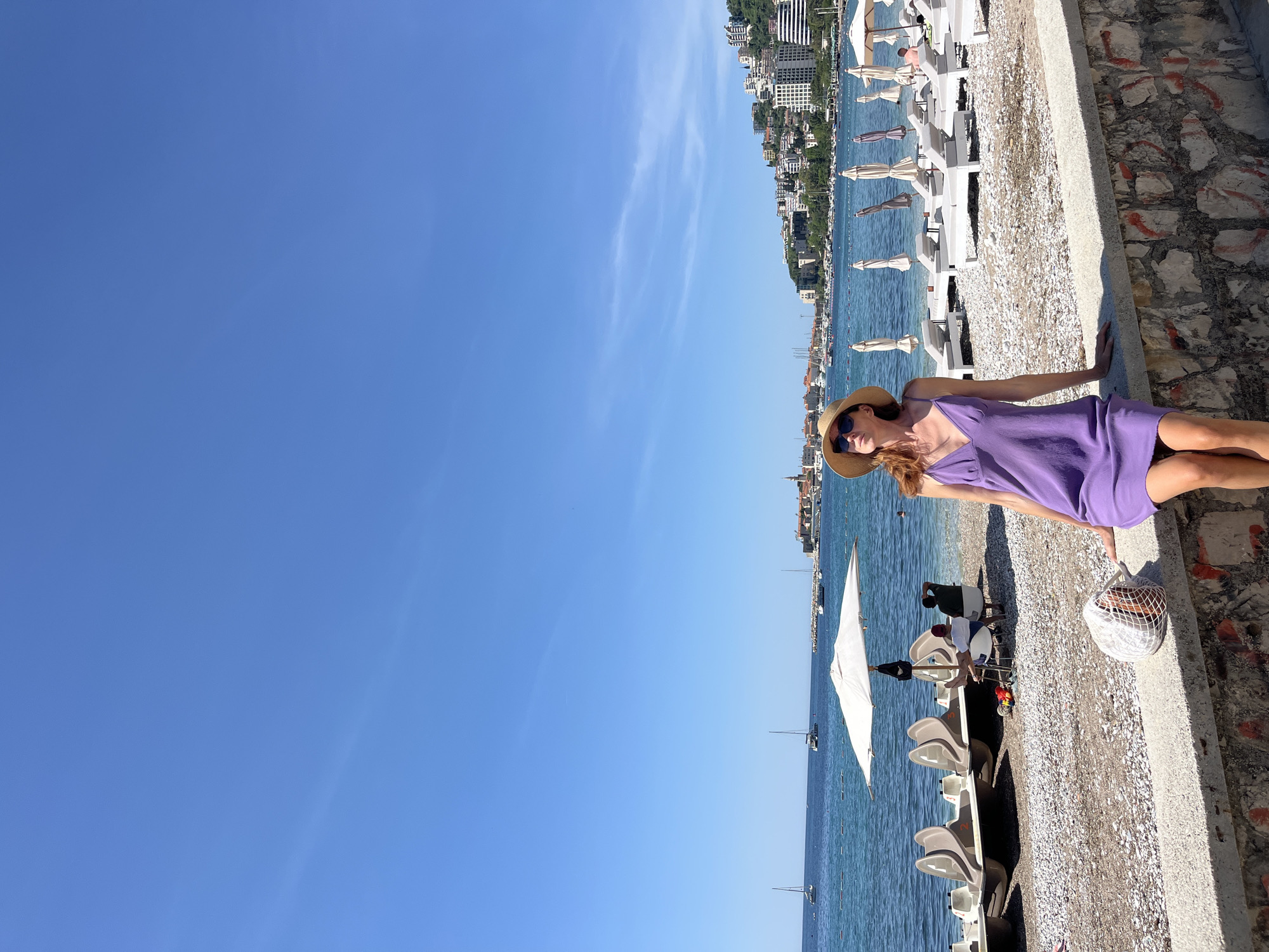 A horizontal, eye-level shot shows a woman on a beach in Budva, Montenegro. The woman, wearing a purple sundress, straw hat, and sunglasses, lies on a concrete barrier alongside a pebble beach. To her right is a white woven bag. The beach features rows of white lounge chairs and umbrellas, and several people are visible attending to watercraft. Beyond the beach is the Adriatic Sea, dotted with sailboats, and in the distance are city buildings and surrounding terrain of Budva, Montenegro. A cloudless blue sky dominates the left portion of the image.