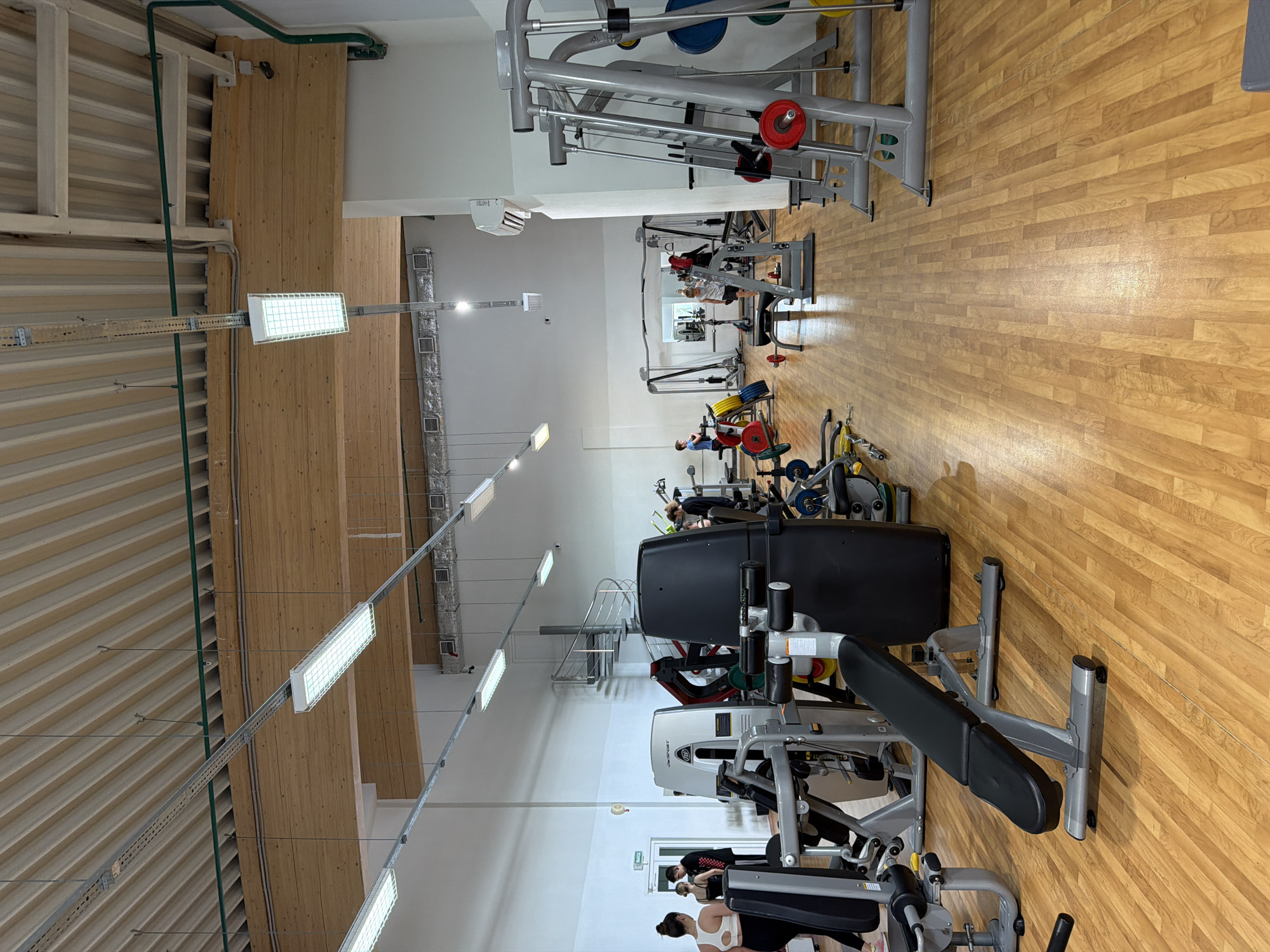 The image shows the interior of a well-lit, modern gym. The gym features a wooden floor and a high ceiling with exposed wooden beams and modern fluorescent lighting. Various exercise machines, including weight machines and benches, are arranged throughout the space. Several people are visible, engaged in activities such as working out or walking. In the background, a spiral staircase leads to an upper level. The environment appears clean and well-maintained, indicative of a functional fitness facility. The gym is located in Podkletnoe, Russia.