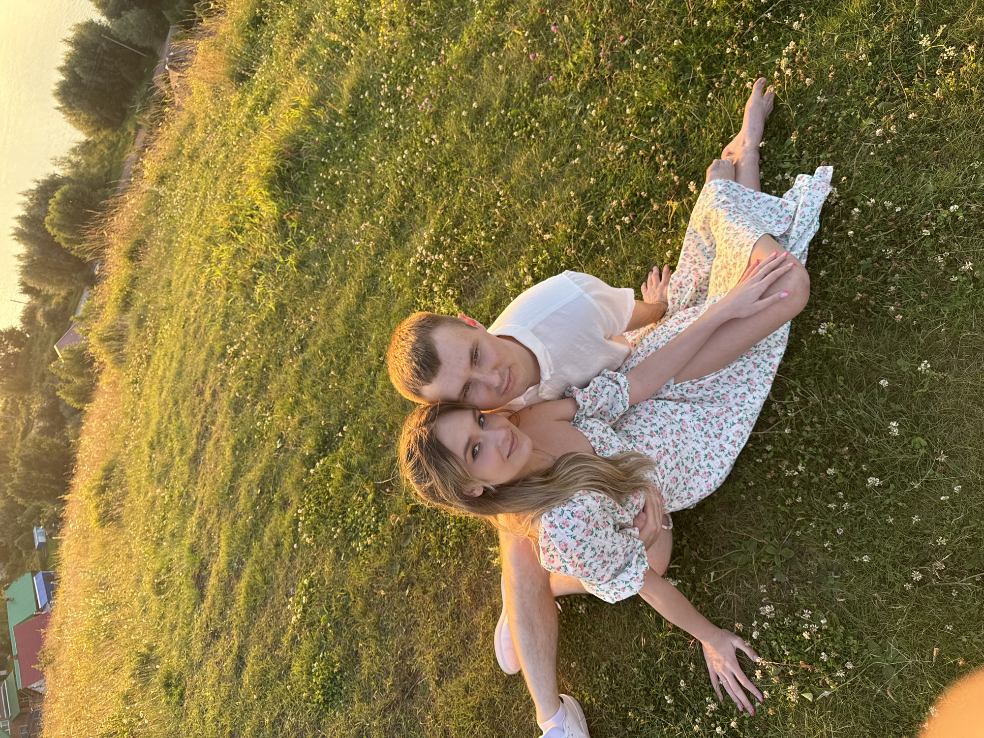 In Olkhovka, Russia, a young couple is captured in a romantic embrace on a grassy slope bathed in the warm glow of the setting sun. The woman, with her long blonde hair cascading around her shoulders, wears a white floral print dress and rests her head on the man's shoulder. The man, dressed in a white t-shirt, has his arm around her. Their legs are intertwined as they sit on the ground, surrounded by small white and pink wildflowers. The background reveals a gentle slope leading to trees and a body of water, with hints of houses and buildings in the distance. The golden hour lighting creates a serene and picturesque atmosphere.