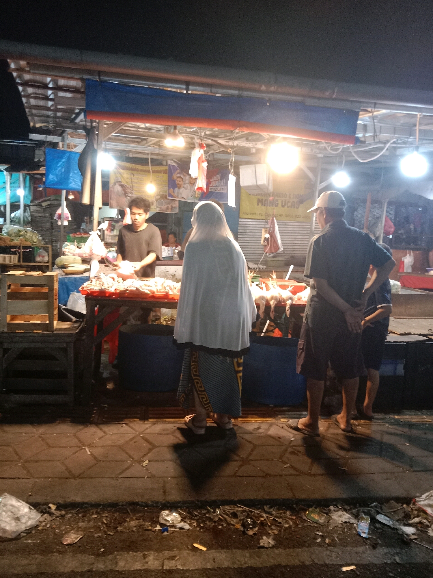 The image depicts a night scene at a street food market, likely located in South Tangerang, Indonesia. In the foreground, a person wearing a white hijab and a patterned skirt stands with their back to the camera, facing a food stall. The stall, illuminated by overhead lights, displays several whole chickens hanging and laid out on a table, alongside other food items. A vendor, a young man in a dark t-shirt, is actively engaged at the stall, handling food. To the right of the vendor, a man wearing a cap and dark clothing stands with his back to the camera, observing the stall. Behind him, another person in a dark shirt and shorts is visible. The ground is paved with square tiles, and the area appears to be a walkway adjacent to the market stalls. Debris and trash are scattered along the edge of the walkway. The overall atmosphere suggests a busy local market at night.