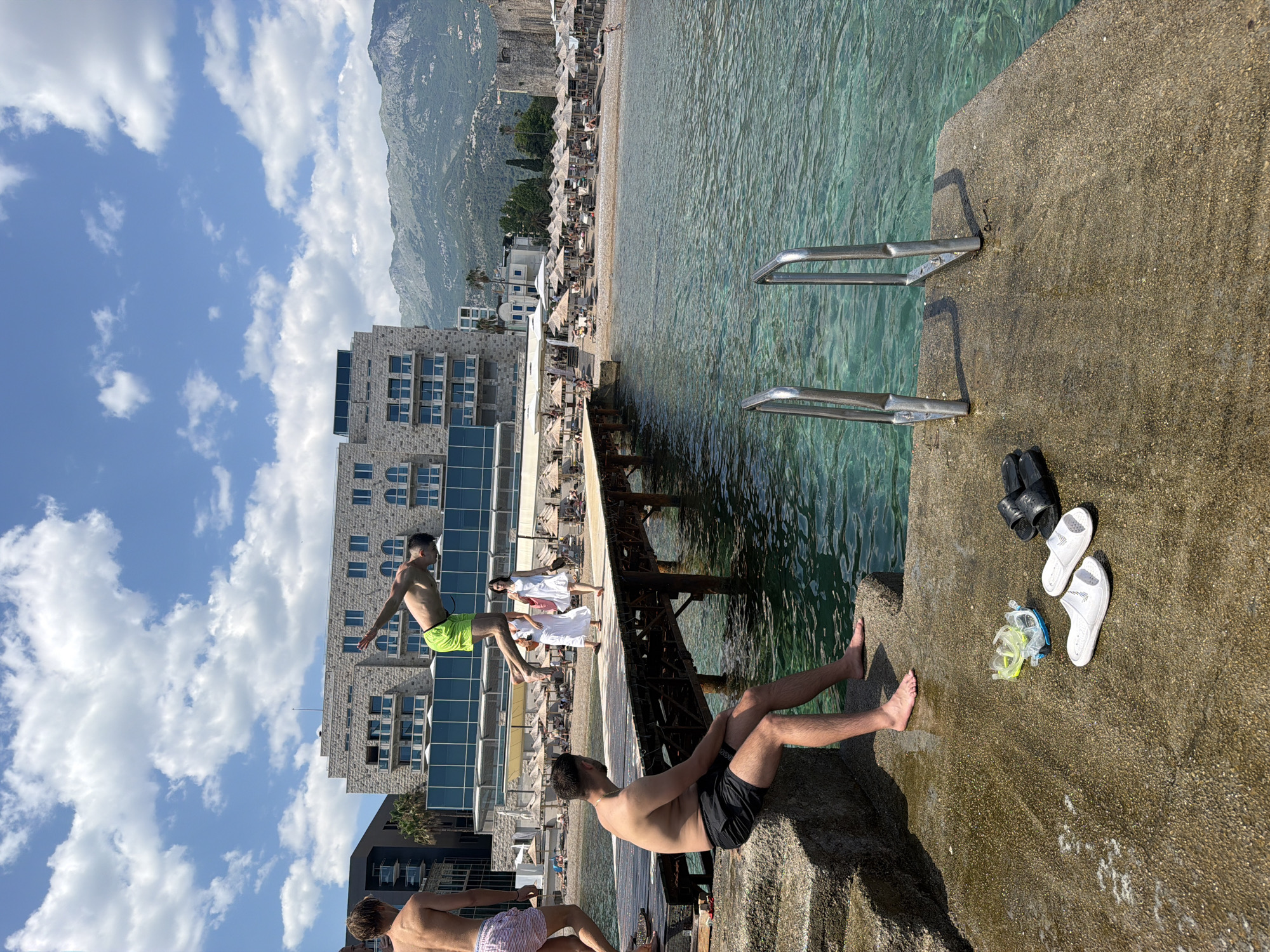 The image depicts a sunny day at a beach resort in Budva, Montenegro. The scene is centered around a concrete pier extending into the clear, turquoise water. On the pier, several people are visible. In the foreground, a man in black swim trunks is sitting with his legs outstretched. To his right, a pair of white flip-flops, a black pair of flip-flops, and a snorkel mask are laid out on the concrete. In the middle ground, a person in bright green swim shorts is in mid-air, jumping from the pier into the water. Behind them, two women in white dresses stand near a railing. The background features a long stretch of beach with numerous umbrellas, a town with buildings, and lush green mountains under a bright blue sky with scattered white clouds. The overall atmosphere is one of leisure and recreation. The time of day appears to be midday, given the bright sunlight and strong shadows.