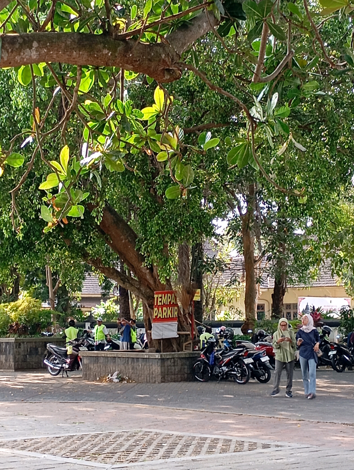 This image depicts an outdoor parking area in Mungkid, Indonesia, under the shade of a large tree. Several motorcycles are parked in a designated lot, with some individuals in yellow vests appearing to be attending to them or the area. Two women, dressed in hijabs and casual attire, are walking across the paved surface. A red sign reading "TEMPAT PARKIR" (Parking Area) is visible on the trunk of the tree. The background shows buildings with tiled roofs, suggesting it's a public or commercial space. The overall scene is illuminated by natural daylight.