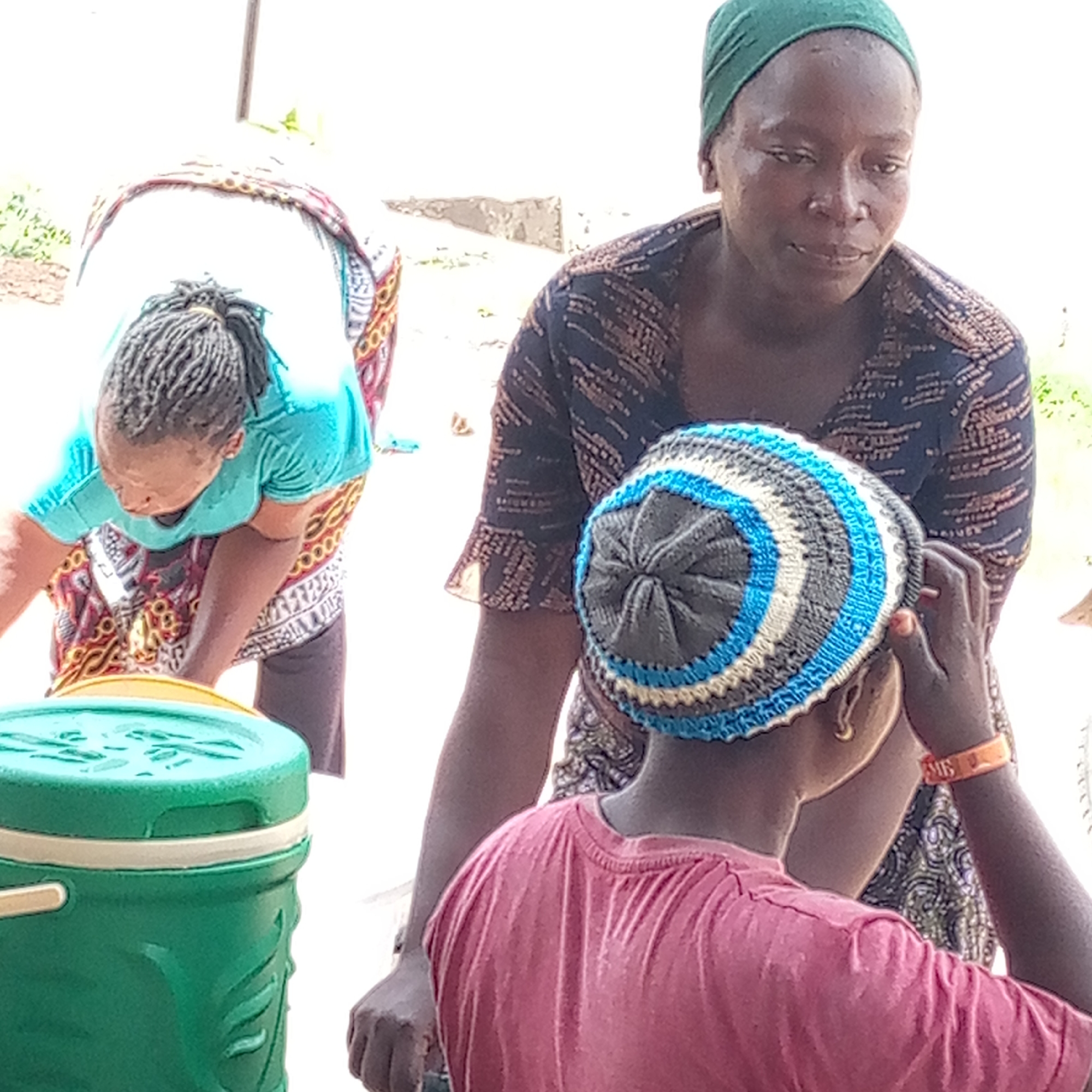 The image shows three women outside. One woman in a green headscarf and a patterned top is looking down at a second woman in a blue and gray knitted hat and a pink shirt. The second woman is turned away from the camera and appears to be touching her head. The third woman is bent over a green and yellow cooler. The background is out of focus. The location is Mararaba, Nigeria.