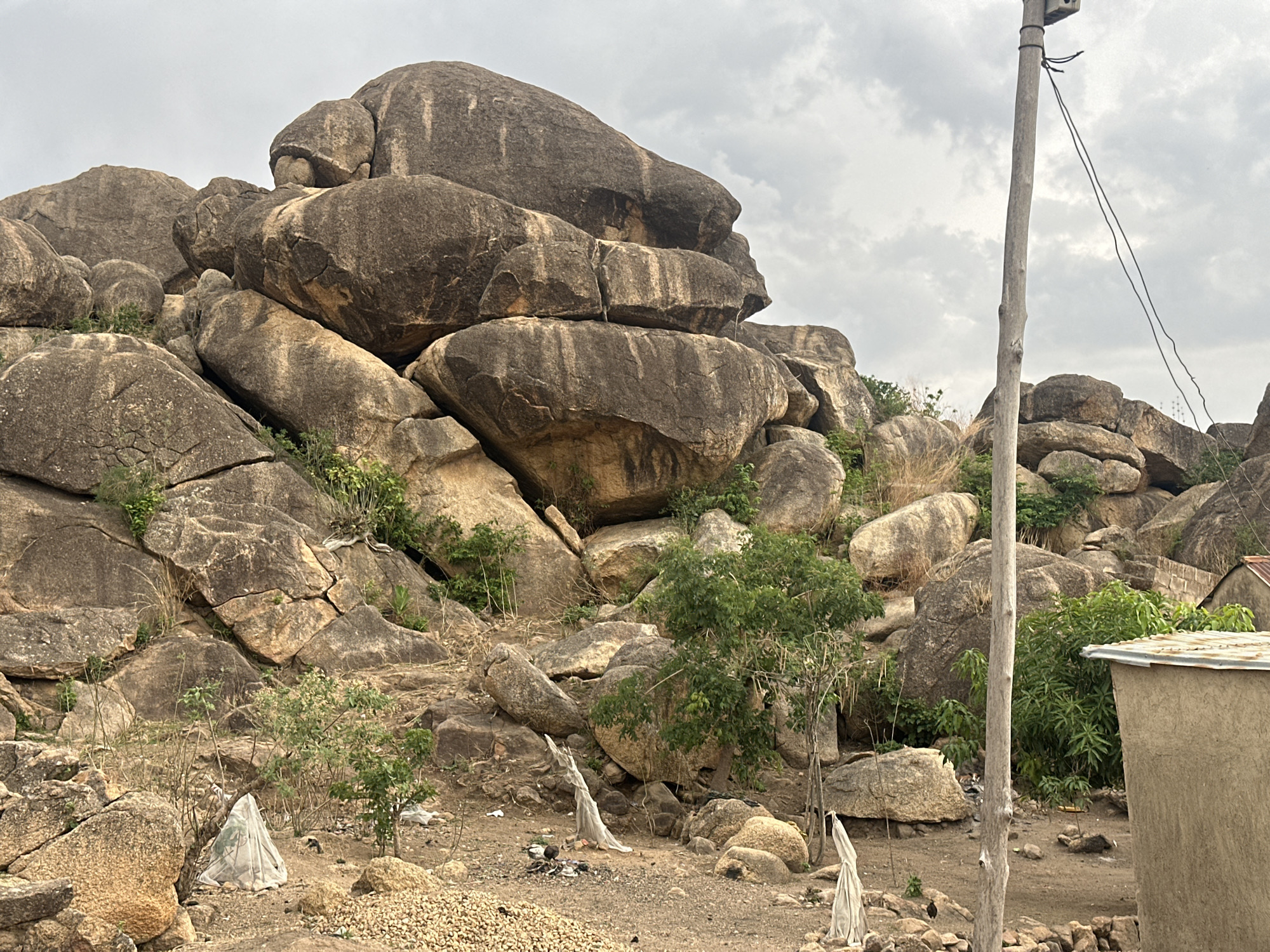The image depicts a rocky landscape with large boulders stacked upon each other. Sparse vegetation, including trees and shrubs, grows amongst the rocks. In the foreground, there are piles of small stones, likely gravel, along with some litter and plastic sheeting covering small plants. To the right, a section of a mud-brick building is visible, and a wooden utility pole stands tall with wires extending from it. The background features more rock formations under an overcast sky. The scene suggests a location on the outskirts of a settlement, possibly in the city of Jos, Nigeria, where such rocky terrain is common. The presence of the building and utility pole indicates human habitation in the area.