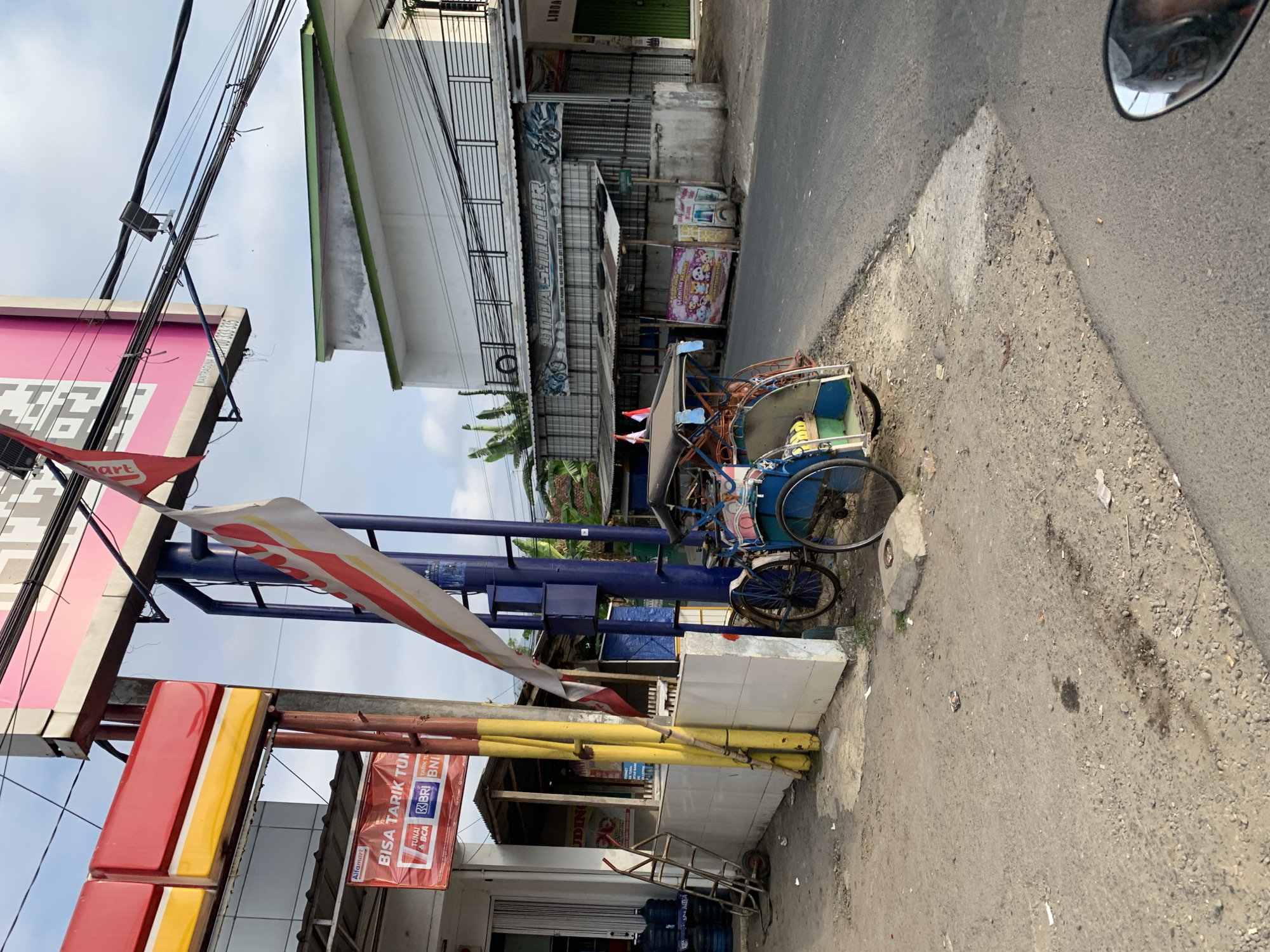 This image captures a street scene in Majenang, Indonesia, near a gas station. A red and yellow sign is visible above the gas pumps, with a red flag hanging from a pole in front of it. To the left of the gas station, a cart is parked next to a white tiled wall, with several large blue water jugs stacked behind it. A metal ladder stands against the wall. In the foreground, a blue and yellow tricycle is parked on the side of the road, with two small flags on its roof. Beyond the tricycle, a row of shops lines the street, including a "DIDA SUMOTOR" shop with a metal shutter. The street is paved, and the sky is overcast. The overall impression is of a busy commercial area with a mix of modern businesses and traditional transportation.