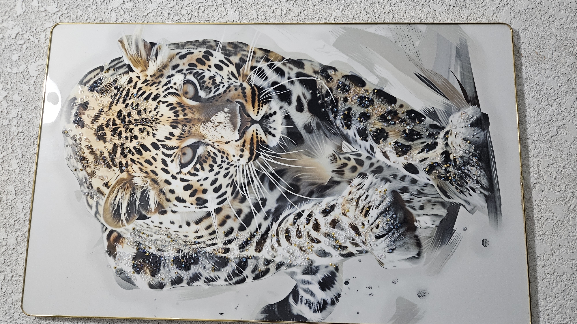 A framed artwork of a leopard is displayed on a textured wall. The leopard is depicted in a close-up, frontal view, with its front paws extended forward as if in motion. The artwork features a realistic portrayal of the leopard's spotted coat, with areas embellished with small, sparkling beads that catch the light. The background of the artwork is a light, neutral color, with subtle brushstrokes creating a sense of depth and movement. The frame is thin and metallic, with a golden hue. The wall behind the artwork has a rough, stucco-like texture.