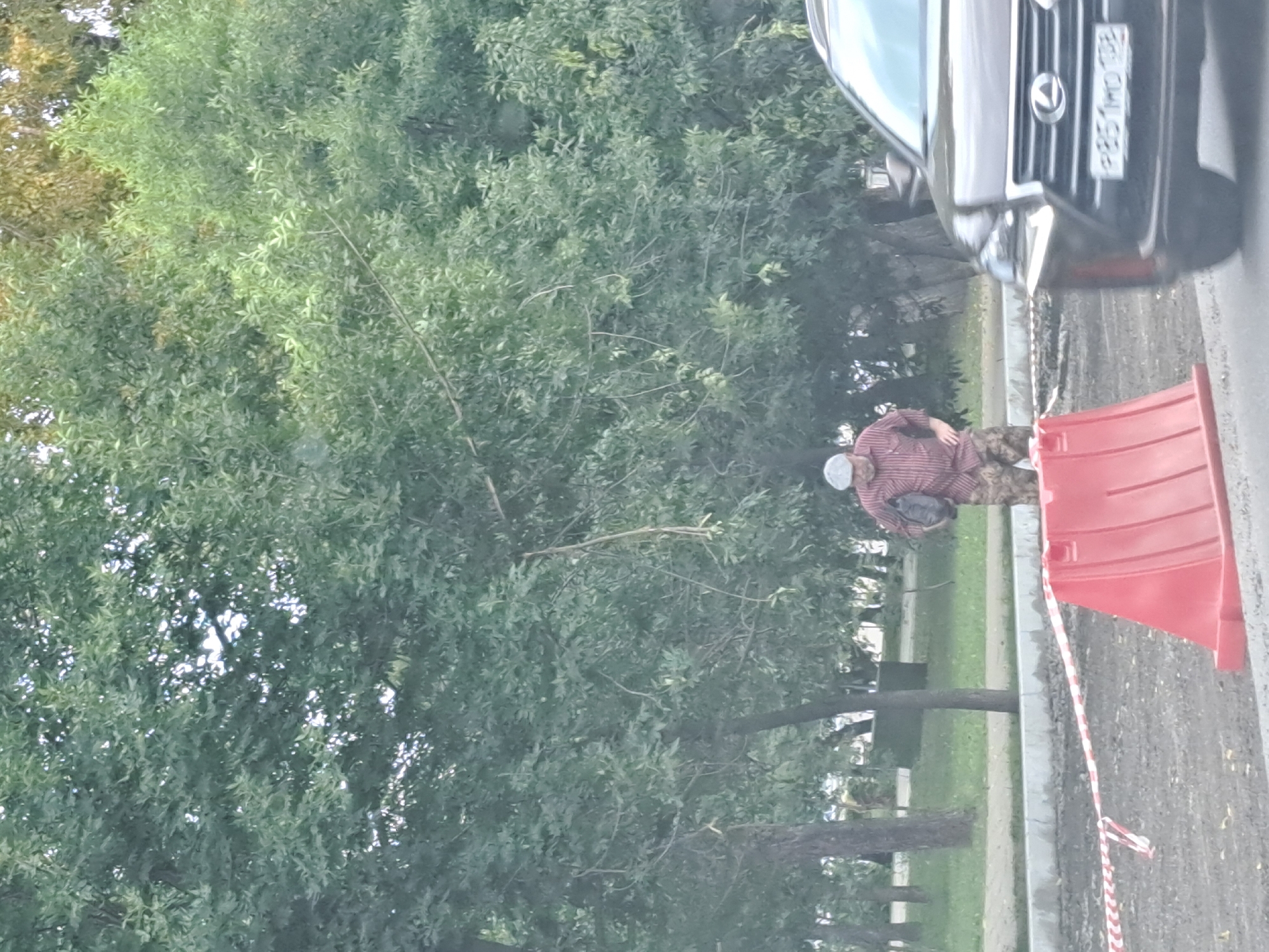 The image shows a man wearing a plaid shirt and camouflage pants, holding a dark bag. He is standing near a construction zone with red traffic barriers and caution tape. A dark-colored Lexus SUV is parked in the foreground. The background consists of trees and foliage. The license plate on the Lexus appears to be "P851MD 138", which is consistent with Russian license plates. While the image itself does not definitively place the scene in Irkutsk, Russia, the license plate format suggests a Russian location.