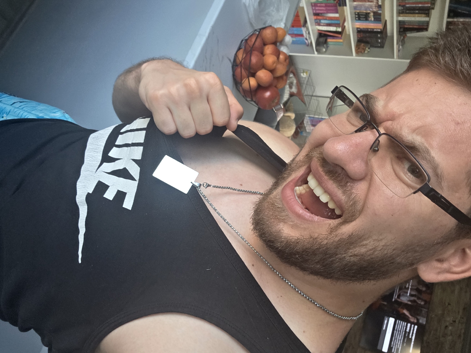 This is an indoor, eye-level, close-up photograph taken in Yagodovo, Bulgaria. The central subject is a Caucasian male, approximately 25-35 years old, with short brown hair, a full beard, and dark-rimmed glasses. He is depicted with an open-mouthed, expressive grimace, showing his upper and lower teeth and tongue, with his eyebrows furrowed. He wears a black tank top with the white "NIKE" logo on the chest and blue patterned shorts, visible at the bottom left. A silver-colored bead chain necklace with a rectangular silver pendant is visible around his neck. His right hand is pulling at the left strap of his tank top. In the background on the left, a white bookshelf filled with numerous multi-colored books is present. On the right, a television screen displays a scene with multiple individuals. On a white countertop in the mid-ground, a black wire bowl contains a collection of red apples and oranges. Behind the countertop, a metal wire rack is discernible.