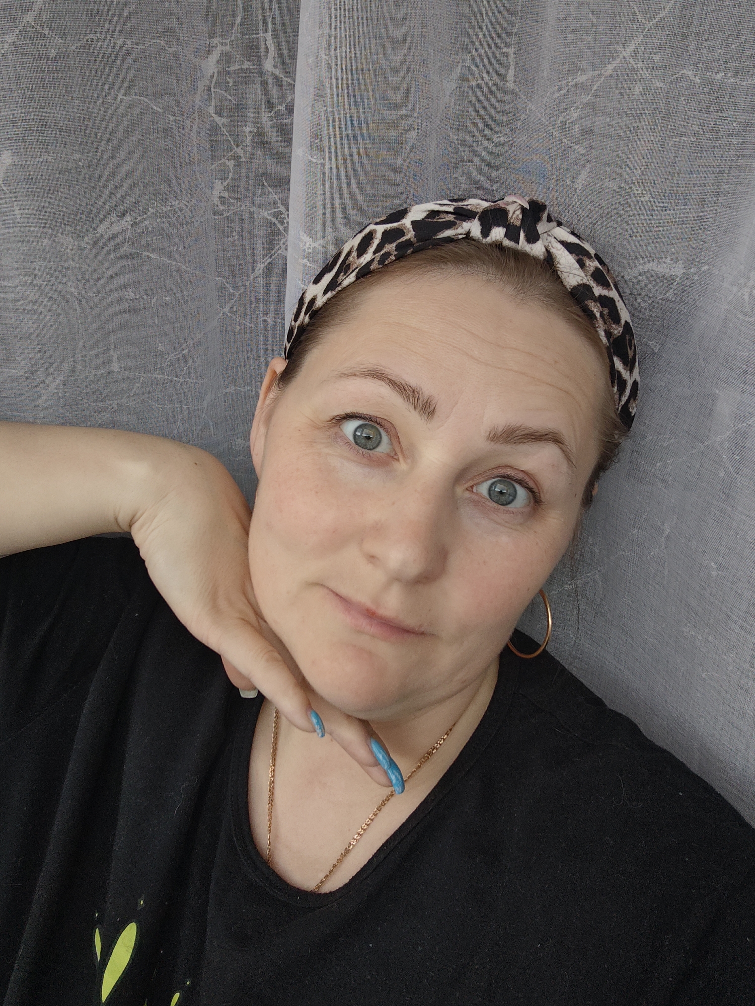 A close-up, eye-level portrait captures a woman indoors, posing for a selfie. She has light skin, striking blue-grey eyes, and light brown eyebrows. Her hair is pulled back, revealing a leopard print headband with a knotted detail at the top. She wears a black t-shirt, a thin gold chain necklace, and a single gold hoop earring is visible on her right ear. Her nails are painted a vibrant light blue, and one hand is positioned against her chin/jawline, fingers slightly spread.
The woman's gaze is directed towards the camera, with a wide-eyed, slightly surprised or curious expression. Her lips are parted in a subtle, soft smile. The background consists of a light grey, semi-sheer curtain with a subtle, irregular texture or pattern, suggesting an indoor setting. There are no external location cues, visible text, or indications of the time of day or weather within the image itself.