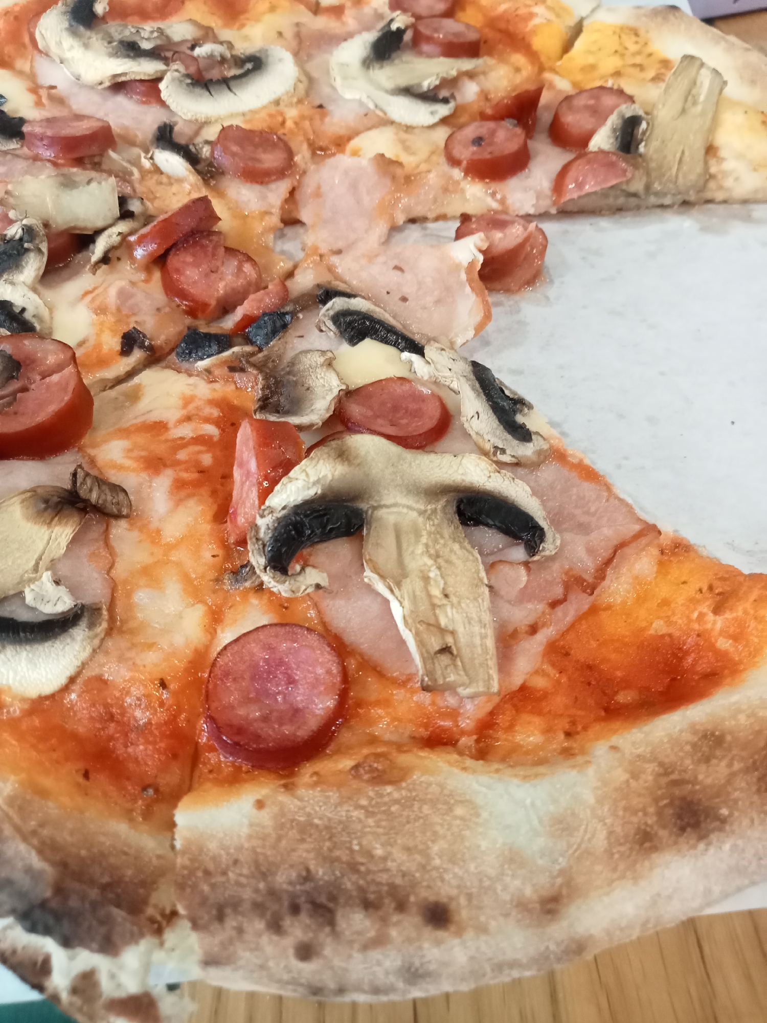 This is a close-up, overhead shot of a pizza with mushrooms and sliced sausages. The pizza is on a white surface, and a portion of it has been removed. The crust is golden brown with some darker, charred spots. The toppings are spread generously across the pizza, with slices of white mushrooms and bright red, circular pieces of sausage. Some pieces of ham are also visible underneath the other toppings. The cheese appears melted and slightly browned in places. The lighting is bright, suggesting an indoor setting, possibly a restaurant or home. There are no people visible in the image, and no text is present. The focus is entirely on the pizza itself, highlighting its texture and ingredients.