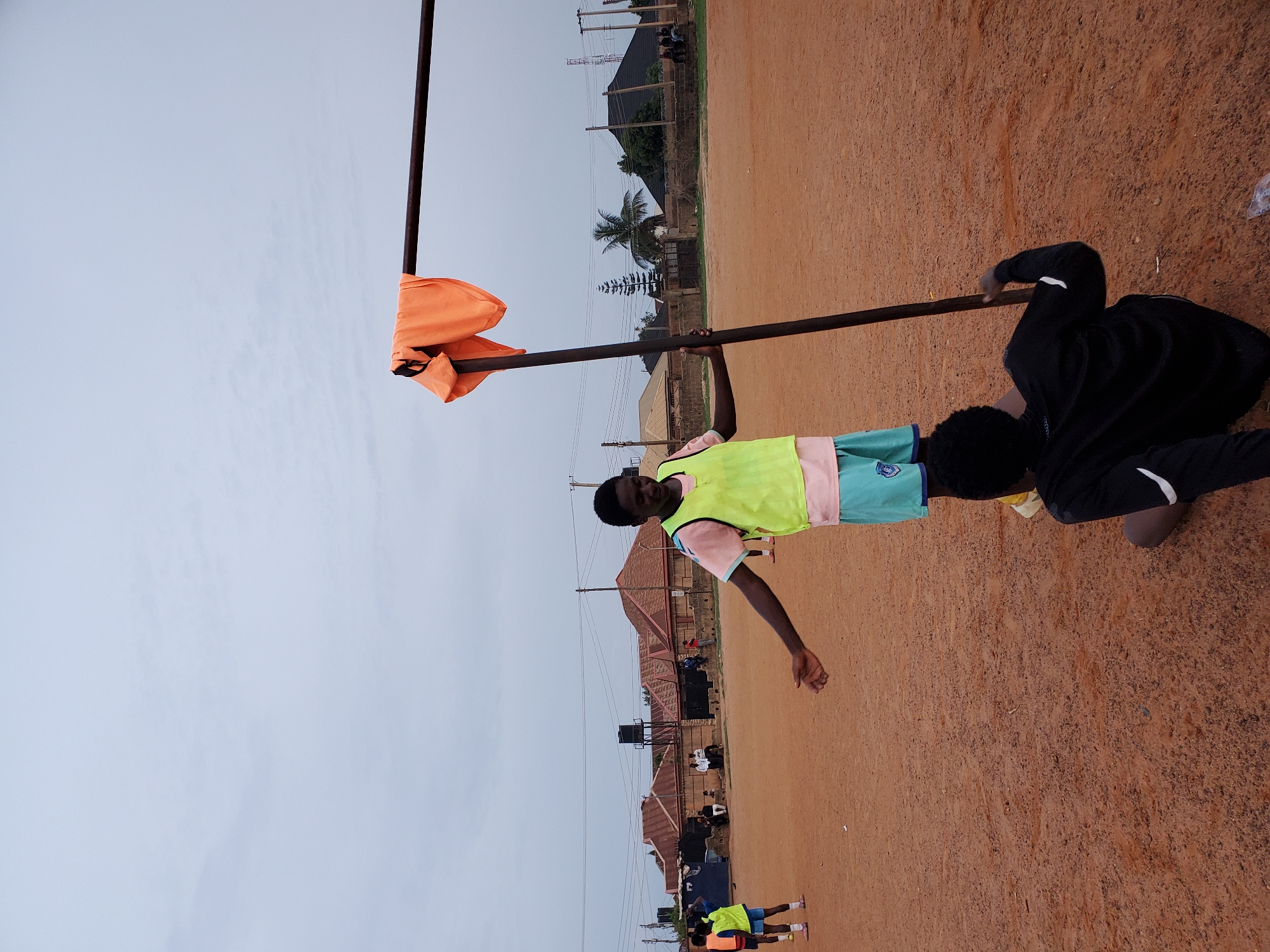 Two young men stand on a dirt field, one holding a metal pole with an orange flag hanging from it. The field is located in Jos, Nigeria. The first man, dressed in a green vest and shorts, holds the pole aloft. The second man, in black clothing, appears to be guiding the pole. In the background are buildings and power lines. Several people stand near buildings in the background.
