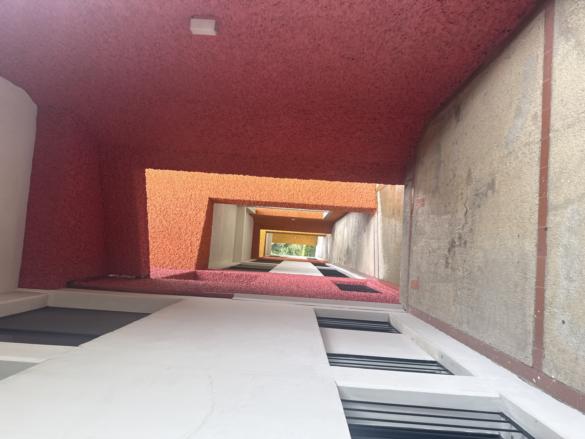 Looking up from an atrium in San Andrés Cholula, Mexico, the view is framed by textured, reddish-pink walls overhead, transitioning to orange further up. Below, a white building facade recedes upwards with evenly spaced dark rectangular windows. The right side of the atrium is a mottled gray concrete wall with a vertical red stripe, while the far end reveals green foliage and an opening to the sky.