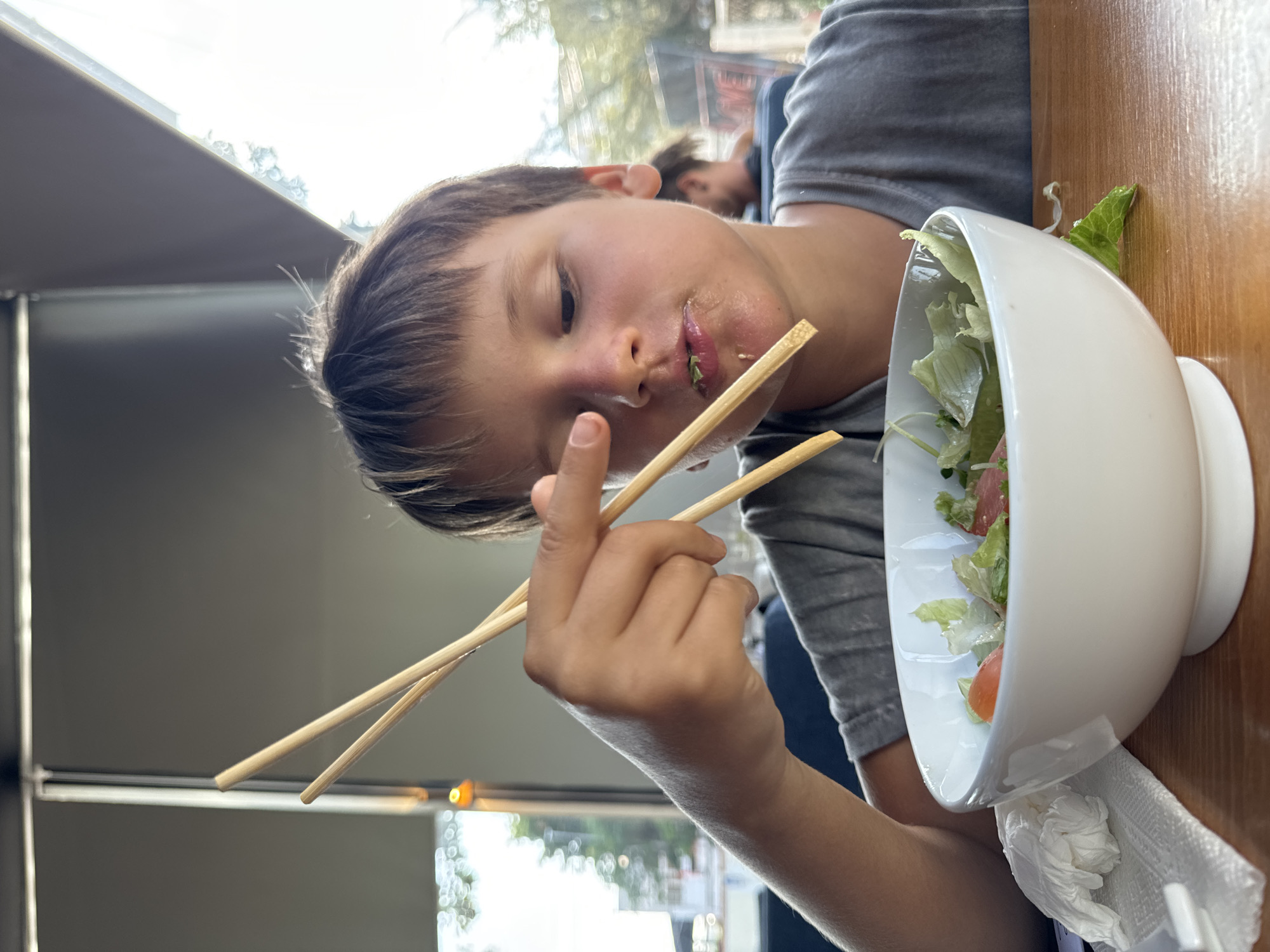 A young boy with brown hair, wearing a grey t-shirt, is seated at a wooden table, eating a salad from a white bowl with chopsticks. He is in the process of bringing a piece of salad to his mouth. A crumpled white napkin is on the table to his left. The background appears to be an indoor-outdoor seating area of a restaurant, with large windows, suggesting a sunny day. Trees and blurred figures are visible in the distance. The scene is set in Phu Thuy Ward, Vietnam.