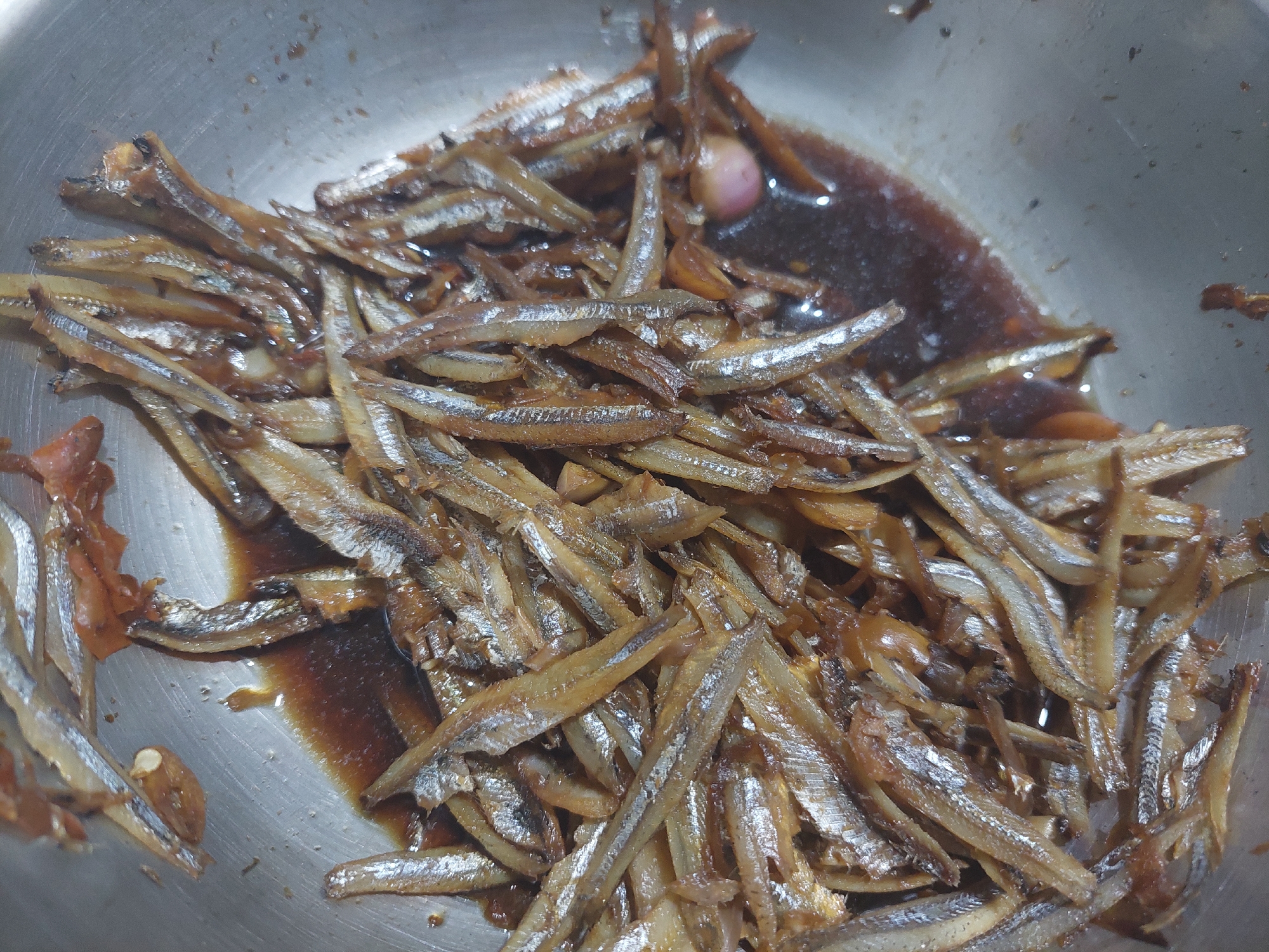 A close-up, overhead shot shows a pile of small, cooked fish in a stainless steel bowl. The fish are a golden-brown color and appear to be coated in a dark, glossy sauce. Some of the fish have visible scales and fins. There are also small pieces of what appear to be garlic or shallots mixed in with the fish. The background is the interior of the metal bowl, which is slightly scratched and reflects light. The image does not contain any specific geographical markers that would allow for its location to be determined within the city of Jelutung, Indonesia.
