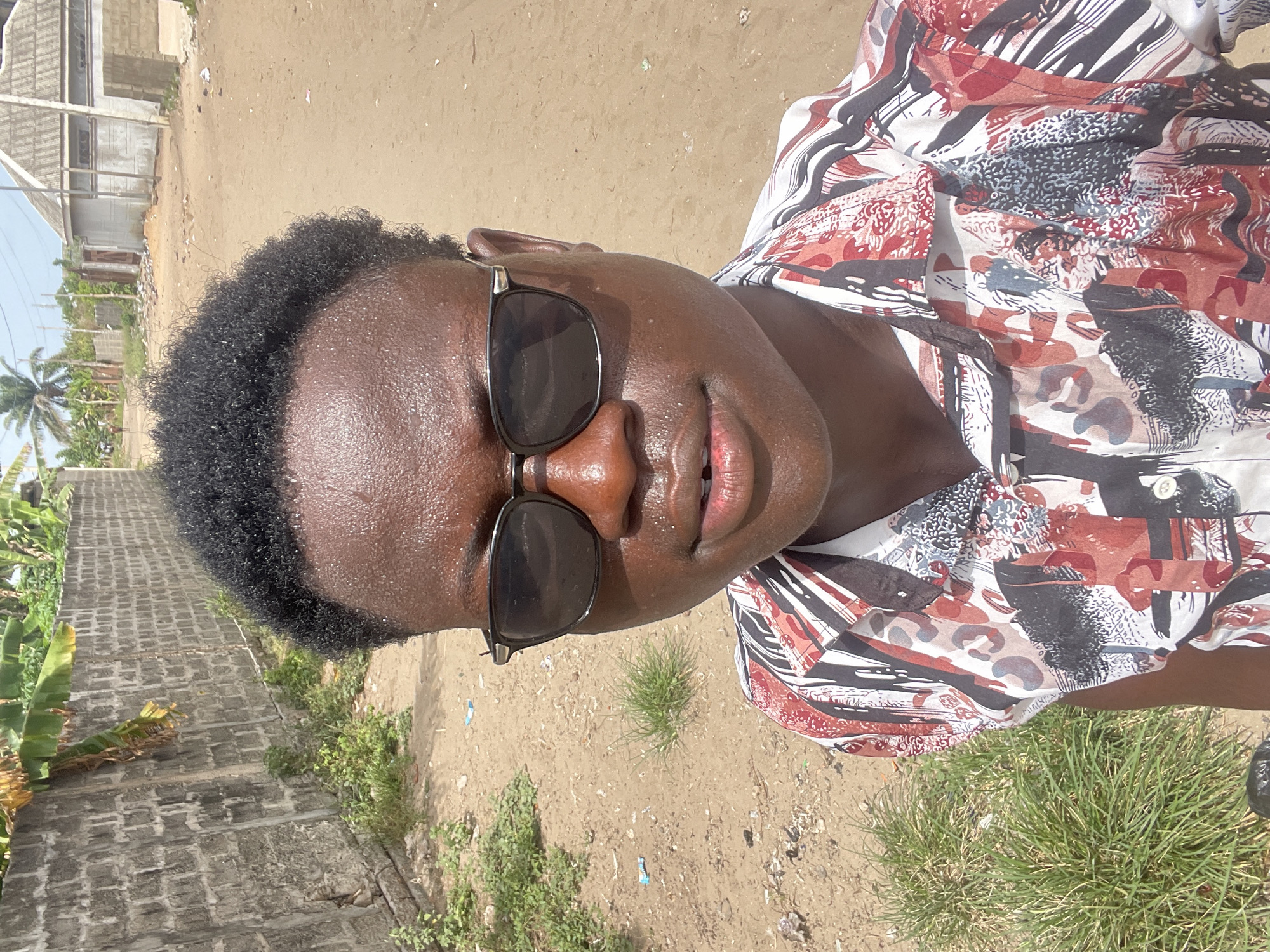 The image shows a dark-skinned person wearing sunglasses and a patterned shirt standing outdoors. The background appears to be a dirt or sandy area with patches of grass and some vegetation. A building or structure is visible in the upper left of the frame. Based on the available context, the person could be located in a less urbanized part of Badagry, Nigeria, with a mixture of natural and built environments. The person is centered and facing the camera and the photograph is taken in daylight.