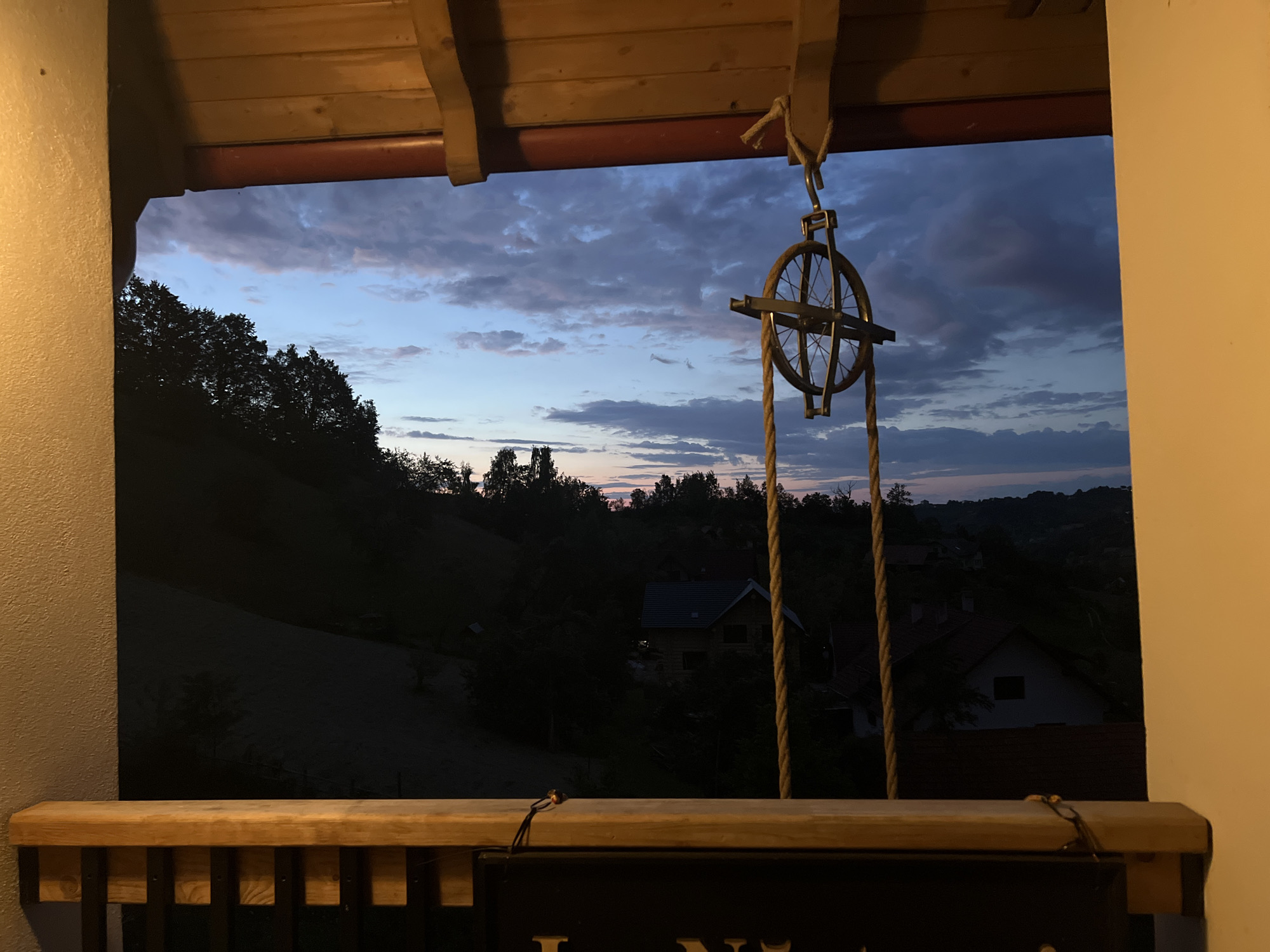 An outdoor scene at dusk viewed through an opening. In the foreground, a wooden railing with dark vertical supports is visible. Above the railing, a wooden beam runs horizontally. Suspended from a metal hook attached to the beam by a rope, a pulley system with a spoked wheel and rope is centered in the frame. The background reveals a landscape with rolling hills covered in trees. Several houses with visible roofs are nestled amongst the trees. The sky displays a mix of dark blue and lighter, pinkish clouds, indicating the end of daylight.