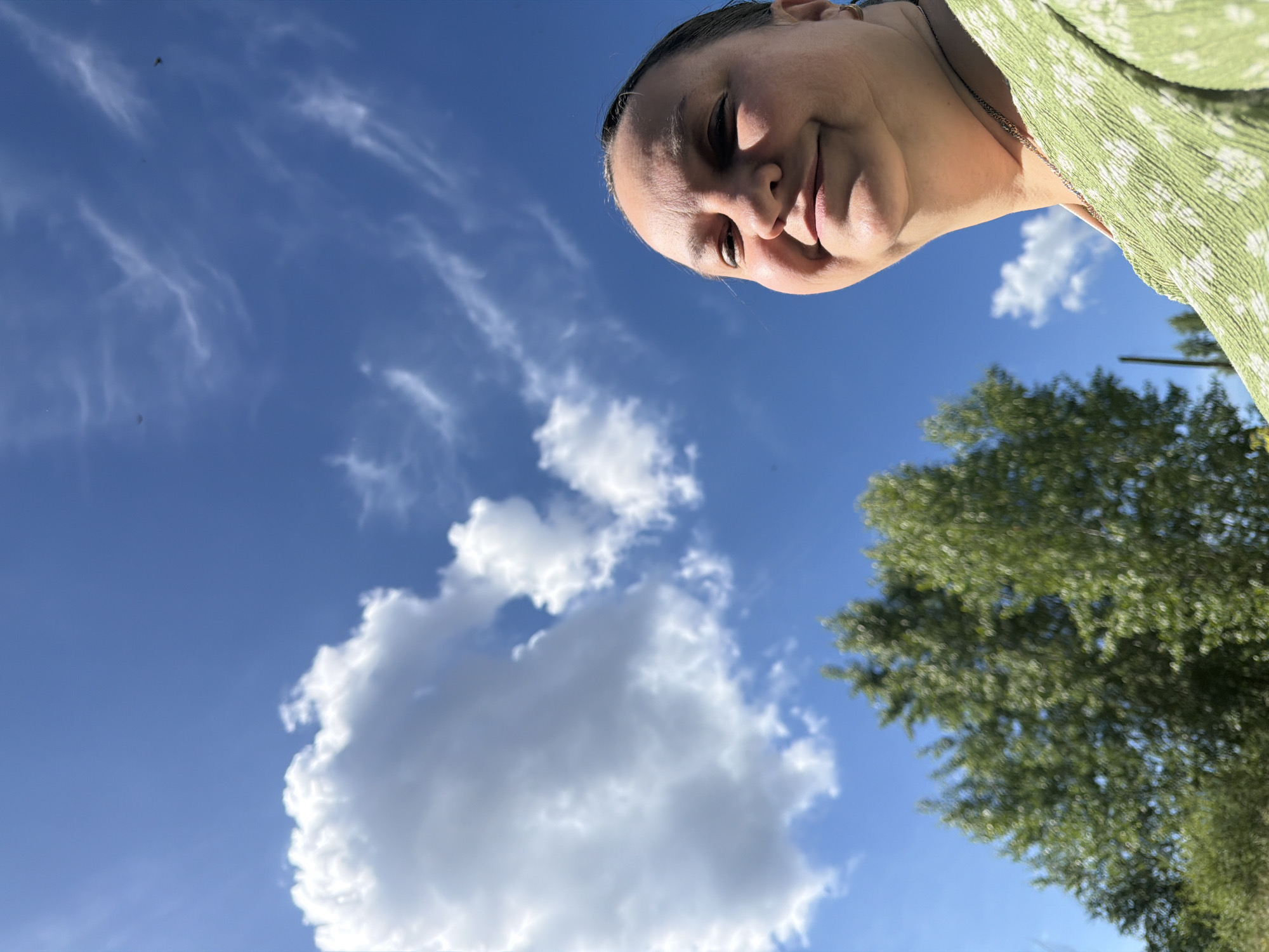 An outdoor, low-angle selfie captures a woman in the foreground, her face turned towards the camera with a slight smile. She has dark hair pulled back, and is wearing a green patterned top and a delicate silver necklace. Behind her, the bright blue sky is dotted with fluffy white clouds and wispy cirrus formations. To the left and lower in the frame, the vibrant green foliage of trees creates a natural backdrop. The image appears to have been taken on a sunny day, likely in a park or natural setting within the city of Chaikovsky, Russia, given the presence of trees and open sky.