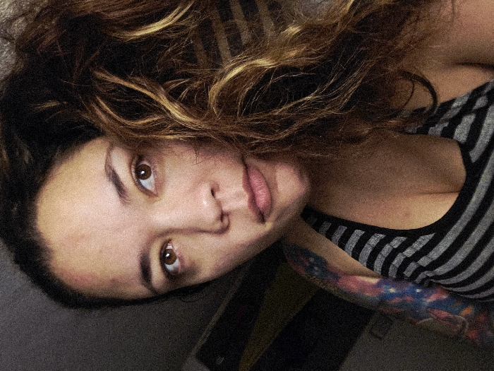 The image displays a female individual in a dimly lit indoor setting, positioned diagonally with her head towards the upper right and her upper torso extending towards the lower left. She has wavy brown hair with lighter streaks, spread across the lower portion of the image and her left shoulder. Her dark brown eyes are open and looking directly at the viewer, and her lips are slightly parted. Observable red marks are present on her forehead, specifically above the left eyebrow. She is wearing a sleeveless top with horizontal black and grey stripes. A multi-colored tattoo, featuring a celestial or cosmic pattern, is visible on her right upper arm. The background is dark and indistinct, offering no discernible environmental features. The precise location within Buenos Aires, Argentina, or any other geographical area, cannot be determined from the visual information presented.