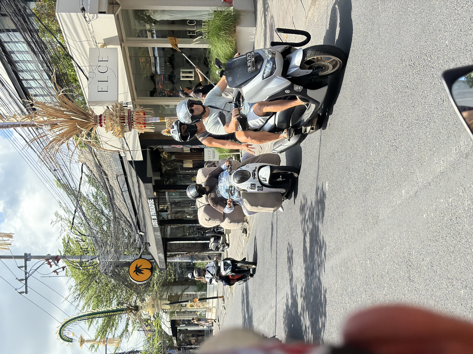 In North Kuta, Indonesia, on a bright, sunny day, a street scene unfolds with multiple people riding scooters. The street is paved, and flanked by shops with awnings and modern building facades. Overhead, electrical wires are densely strung between poles, a common sight in many Indonesian cities. In the foreground, a person's shoulder and the edge of a vehicle's mirror are visible, suggesting the photo is taken from another moving vehicle.
Further down the street, two individuals are on a silver scooter. The driver, a man wearing a light blue t-shirt, white shorts, sunglasses, and a silver helmet, is operating the scooter. A woman behind him, wearing a black tank top and a dark helmet, is a passenger. She is leaning slightly forward. This scooter has a license plate that reads "DK 2076 FCD 06-26".
To their left, another scooter, white with a large cargo area covered in light brown material, is ridden by a person in a brown shirt and dark helmet. This scooter is carrying what appears to be large bales of material strapped to its rear.
Further still, a black scooter is ridden by a person wearing a white shirt and a dark helmet. In the background, other people are seen walking on the sidewalk and shopping. Signage on the buildings includes "LOST IN PARADISE" and "ELCE", indicating retail establishments. Decorative elements, including what appear to be traditional Balinese offerings and palm fronds, are visible, adding to the local context of the scene. The overall impression is of a bustling commercial street in Bali, Indonesia.
