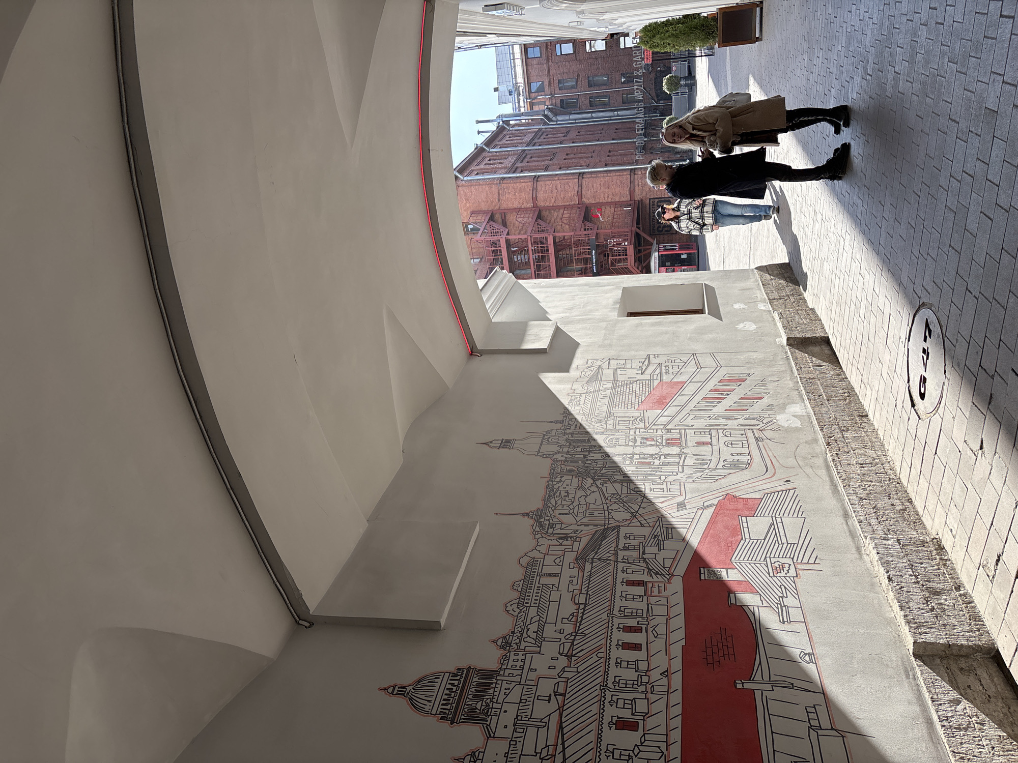 The image depicts a paved urban passageway in Saint Petersburg, Russia, specifically identified as a section within the Berthold Centre (Бертольд Центр), located at Grazhdanskaya St, 13-15. This identification is supported by the distinctive mural portraying the Saint Petersburg skyline, including Saint Isaac's Cathedral, on the left wall, and legible signage in the background for "PROSTRANSTVO" and "PG-00 ERAAGG APZIZ & GARDEN."
The passageway features a light-colored arched ceiling, with a red neon-like strip light running along the inner curve of the archway at its far end. The left wall is adorned with a large mural in black outline and selective red accents, depicting the rooftops and prominent architectural landmarks of Saint Petersburg. Below the mural, the wall transitions to a rough-hewn stone or brick base. The ground is paved with light-colored rectangular pavers, and a circular manhole cover marked "G47" is visible in the foreground. The right wall is plain, light-colored, and partially obscured by a small, potted evergreen plant near the archway's exit.
The scene is illuminated by natural sunlight, casting sharp shadows across the mural, pavement, and individuals. Beyond the archway, an open, brightly lit courtyard is visible, flanked by multi-story red brick buildings featuring numerous windows and external metal staircases or fire escapes.
Three individuals are present. In the sunlit foreground, a male with light-colored hair, dressed in a dark long coat, dark trousers, and boots, stands facing a female. He holds a smartphone in his right hand. The female, with blonde hair, wears a light brown shearling-style coat, dark trousers, and boots, and carries a light-colored bag on her left shoulder; she is looking at the smartphone held by the male. Further back in the passageway, partially in shadow, a third individual walks away from the camera towards the courtyard, wearing a checkered jacket, blue jeans, and a dark cap.