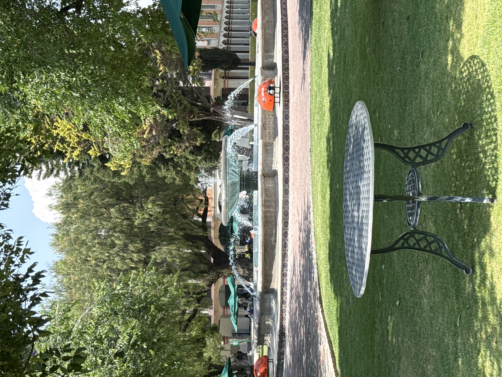 The image depicts a park in San Andrés Cholula, Mexico. A stone fountain with multiple tiers is the focal point, water cascading from each level. Beside the fountain, a decorative orange ladybug statue stands on a tiled walkway that separates the fountain from a grassy area where a round metal table with ornate black legs sits. Lush green trees provide shade and surround the park. In the background, glimpses of buildings with arched windows and green umbrellas hint at a nearby commercial area. People are visible under the umbrellas, suggesting a cafe or outdoor seating area.