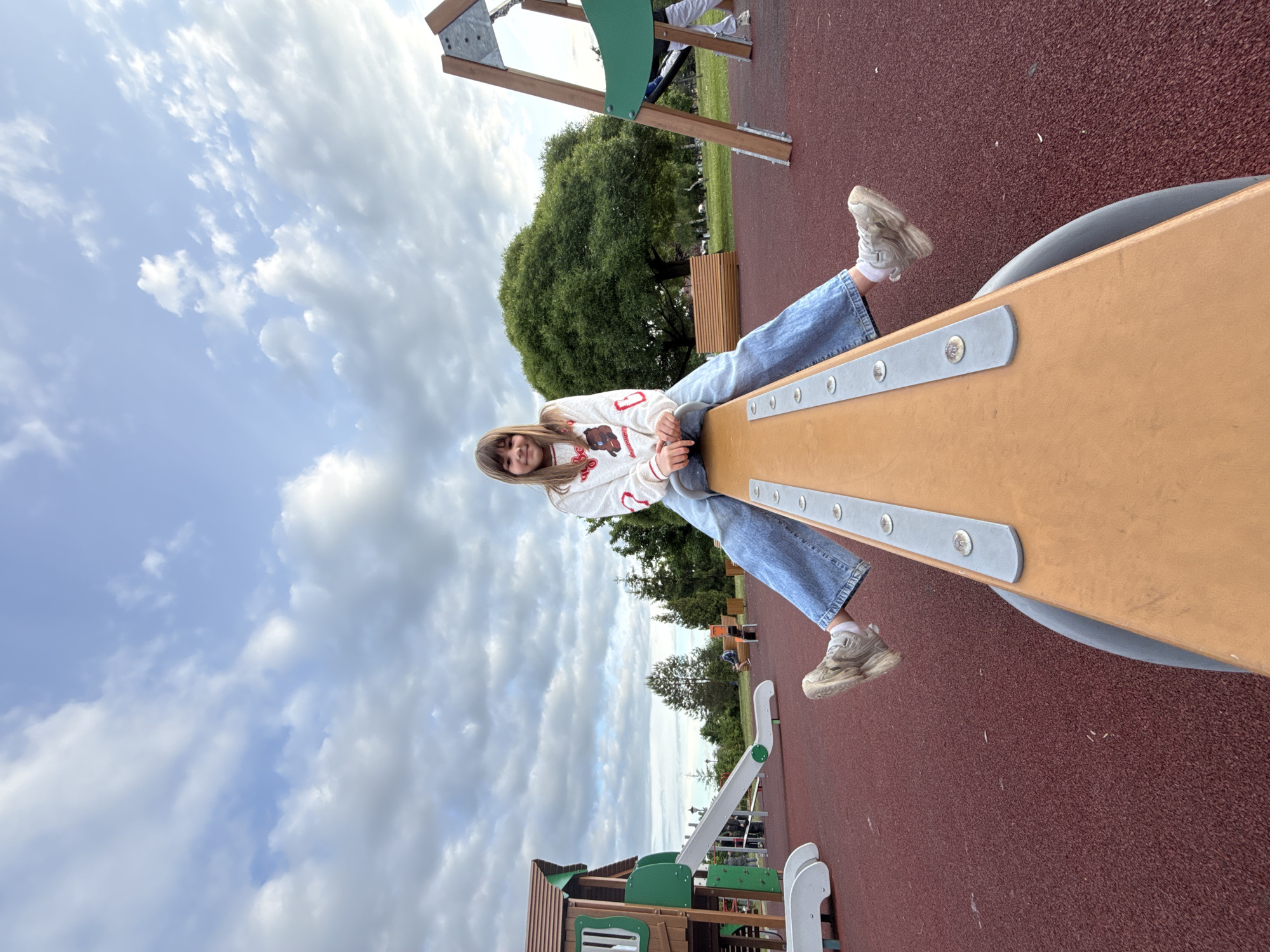 A young female is seated on a seesaw at a playground in Заволжье, Russia. She is positioned on one end of the light brown seesaw, looking towards the camera with a slight smile. She has long, dark blonde hair parted in the middle.
She is wearing a white long-sleeved top with a graphic design, likely an animal or bear head, on the front. Her attire also includes loose-fitting blue jeans, white socks, and light-colored chunky sneakers. Her hands are resting on the seesaw's handles.
The playground features a reddish-brown, textured rubberized ground. In the background, other green and brown playground structures, including a slide and a playhouse, are visible. Lush green trees and a park bench can be seen further back. The sky is partly cloudy with a mix of white and gray clouds against a lighter blue, suggesting daytime. The scene conveys a relaxed and enjoyable atmosphere at a children's play area. No visible text is discernible on the subjects or objects.