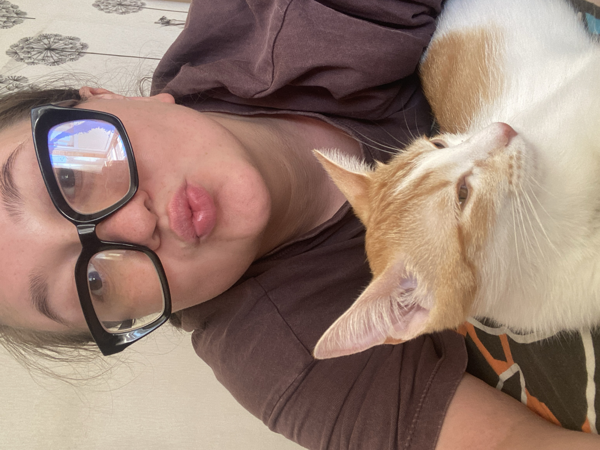 A woman with glasses lies on her back with an orange and white cat on her chest. The woman wears black-framed glasses and a brown shirt. The cat's head rests on the woman's chest. The background appears to be a light-colored surface with a pattern, possibly a tablecloth. The location is not discernible from the image.