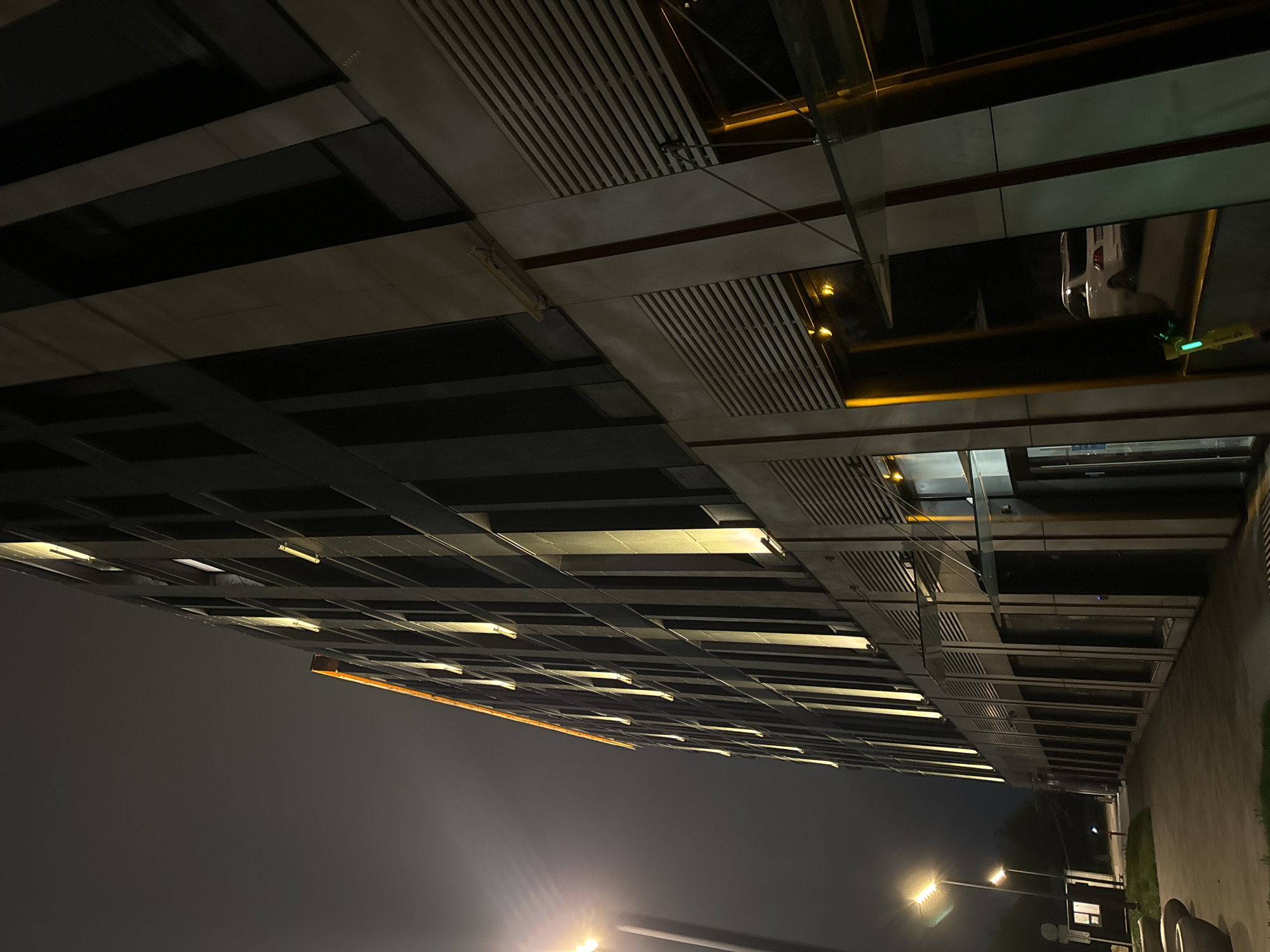 The image depicts an upward-angled view of a modern building exterior at night. The building features a grid-like facade with alternating sections of solid panels and horizontally slatted surfaces. Some sections are illuminated, creating a pattern of light and shadow. The building appears to have a glass overhang or canopy in some areas, reflecting the lights and surrounding environment. At the bottom of the image, streetlights illuminate a section of sidewalk and landscaping adjacent to the building. The location is not identifiable as Moscow, Russia, without additional information.