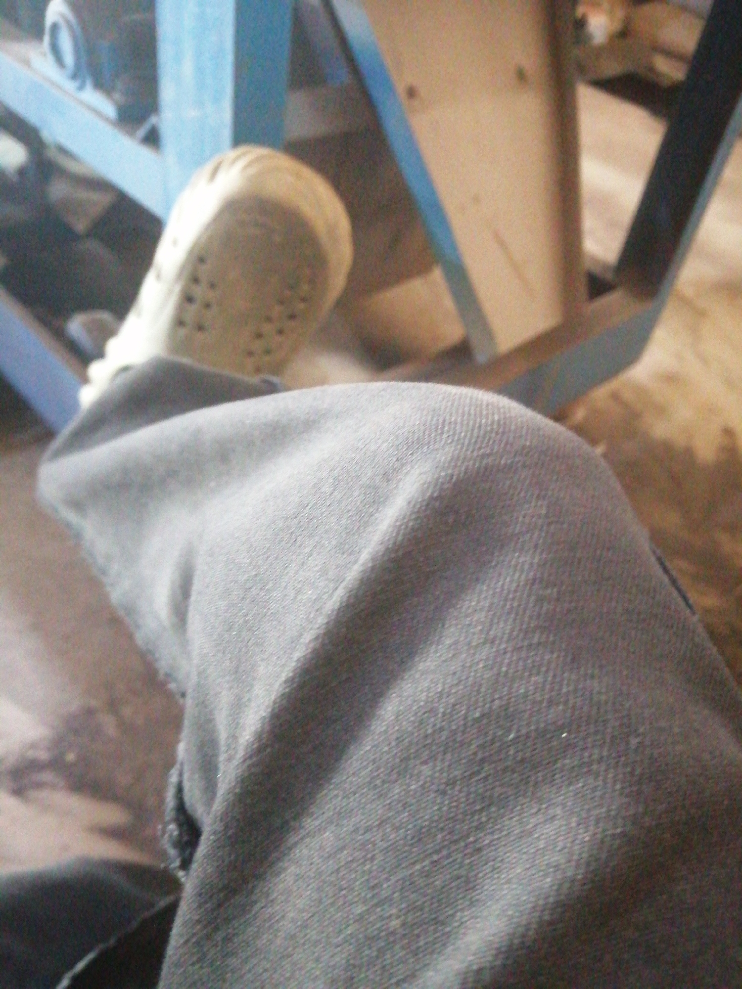 The image displays a close-up of a person's knee and lower leg, covered by dark fabric, likely pants or jeans. A light-colored shoe with a textured sole is visible on the left side of the frame, partially obscuring the leg. In the background, a blue metal structure, possibly part of a machine or scaffolding, is present. The overall scene suggests an indoor or semi-outdoor industrial or workshop setting. No specific landmarks of Jos, Nigeria are identifiable within the frame.