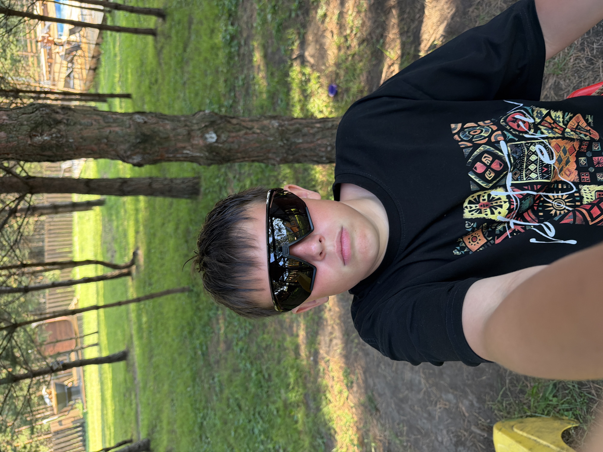 This is a vertical, medium shot of a young person wearing black sunglasses and a black t-shirt with a colorful, geometric pattern on the front. The subject is looking towards the camera, with a neutral expression. The background shows a grassy area with trees and a wooden fence. There are shadows from the trees, suggesting it is daytime. A portion of a yellow object is visible in the lower right corner of the frame. The overall scene appears to be outdoors, possibly a park or a wooded area. The location context provided is Elekmonar, Russia, which suggests a natural, possibly rural or suburban setting. There is no visible text in the image.