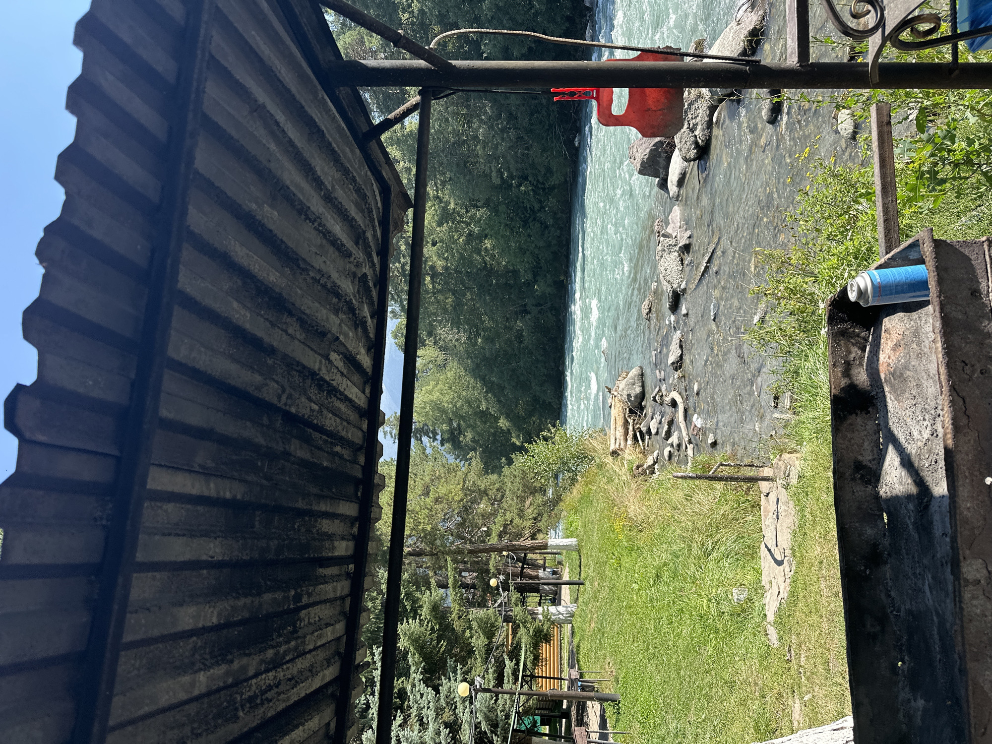The image depicts an outdoor seating area with a metal roof structure overlooking a fast-flowing river in Arkhyz, Russia. In the foreground, a rusty barbecue grill is visible, with a blue gas canister placed inside it. To the right, a red object, possibly a buoy or a piece of equipment, hangs from a metal pole. The river, with its turquoise water, flows over rocky terrain, bordered by lush green grass and dense trees on the opposite bank. Several conifer trees are present on the left side of the scene, along with a wooden structure and lighting fixtures. The sky is clear and blue. The overall impression is of a serene natural setting suitable for outdoor recreation.