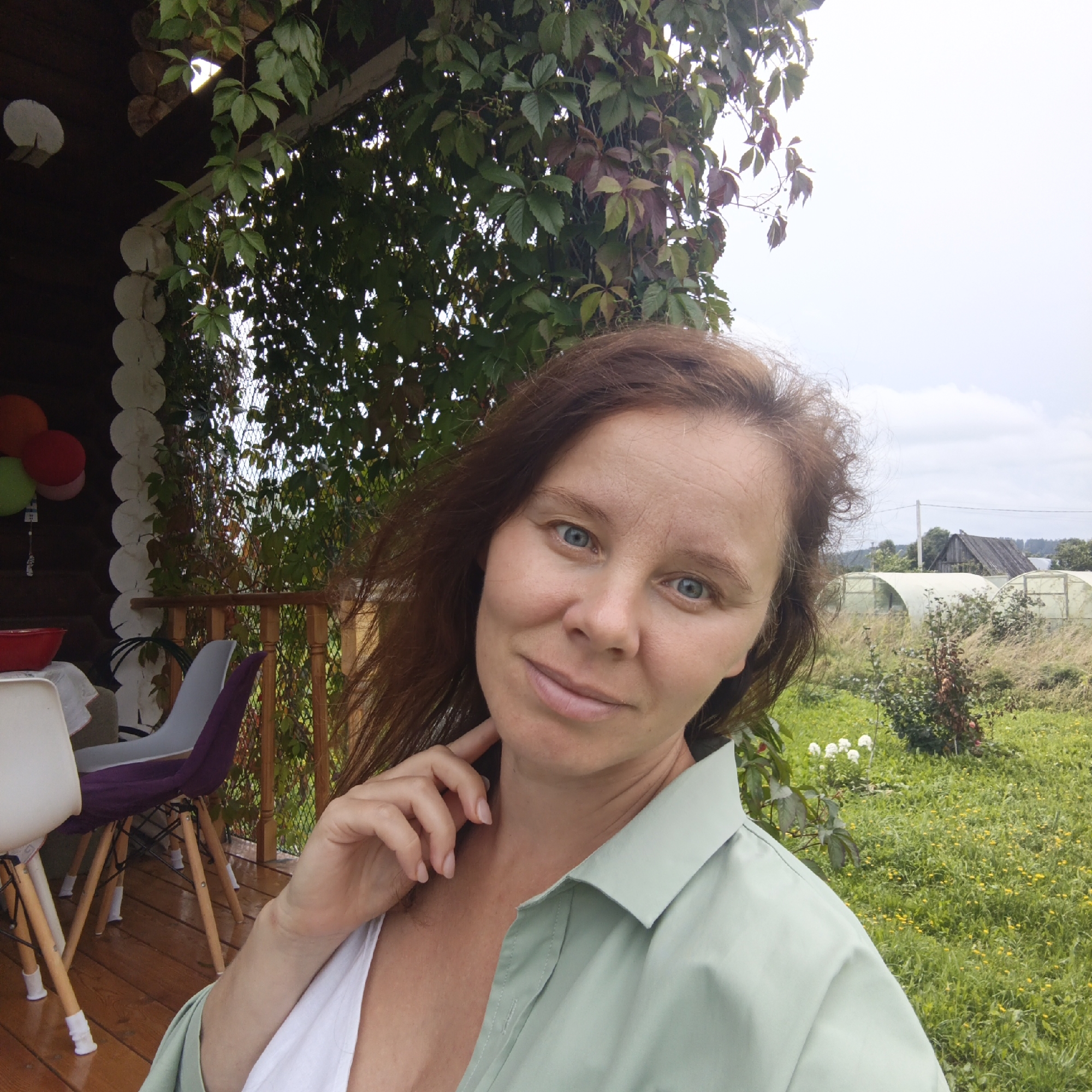The image is a close-up selfie of a woman with brown hair and blue eyes, taken outdoors. She is wearing a light green collared shirt over a white top. Her right hand is lightly touching her neck, and she is looking towards the camera with a slight smile. The background shows greenery, including some vines with red and green leaves climbing on a wooden structure, possibly a porch or veranda of a log house. There are also some colorful balloons visible on the left side of the image. In the further background, there is a field with grass and yellow flowers, some greenhouses, and a wooden building. The overall impression is of a casual, outdoor setting, likely in a rural area. No specific location within the city of Большой Букор, Russia can be determined from the image itself.