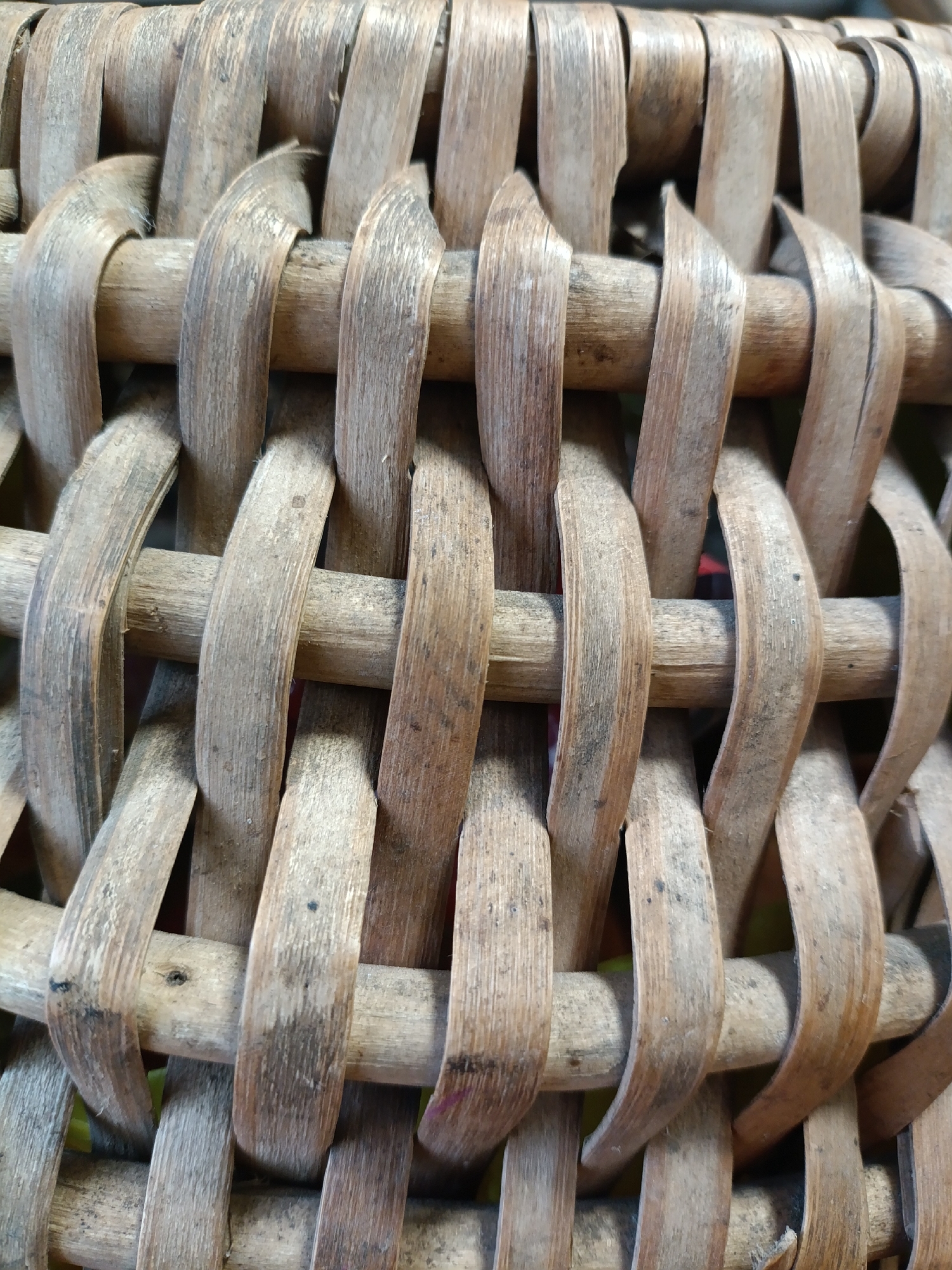 The image shows a close-up, tightly cropped view of a woven basket. The basket appears to be made of natural, dried materials, likely rattan or bamboo, with a light brown, weathered texture. The weaving pattern is visible, with vertical strands passing over and under horizontal strands, creating an interlaced design. The surface of the material shows variations in color and texture, with some areas appearing smoother and lighter, while others are darker and slightly rougher, suggesting natural imperfections and wear. There are no discernible objects, people, or environmental details beyond the basket itself. The lighting is even, highlighting the intricate details of the weave.